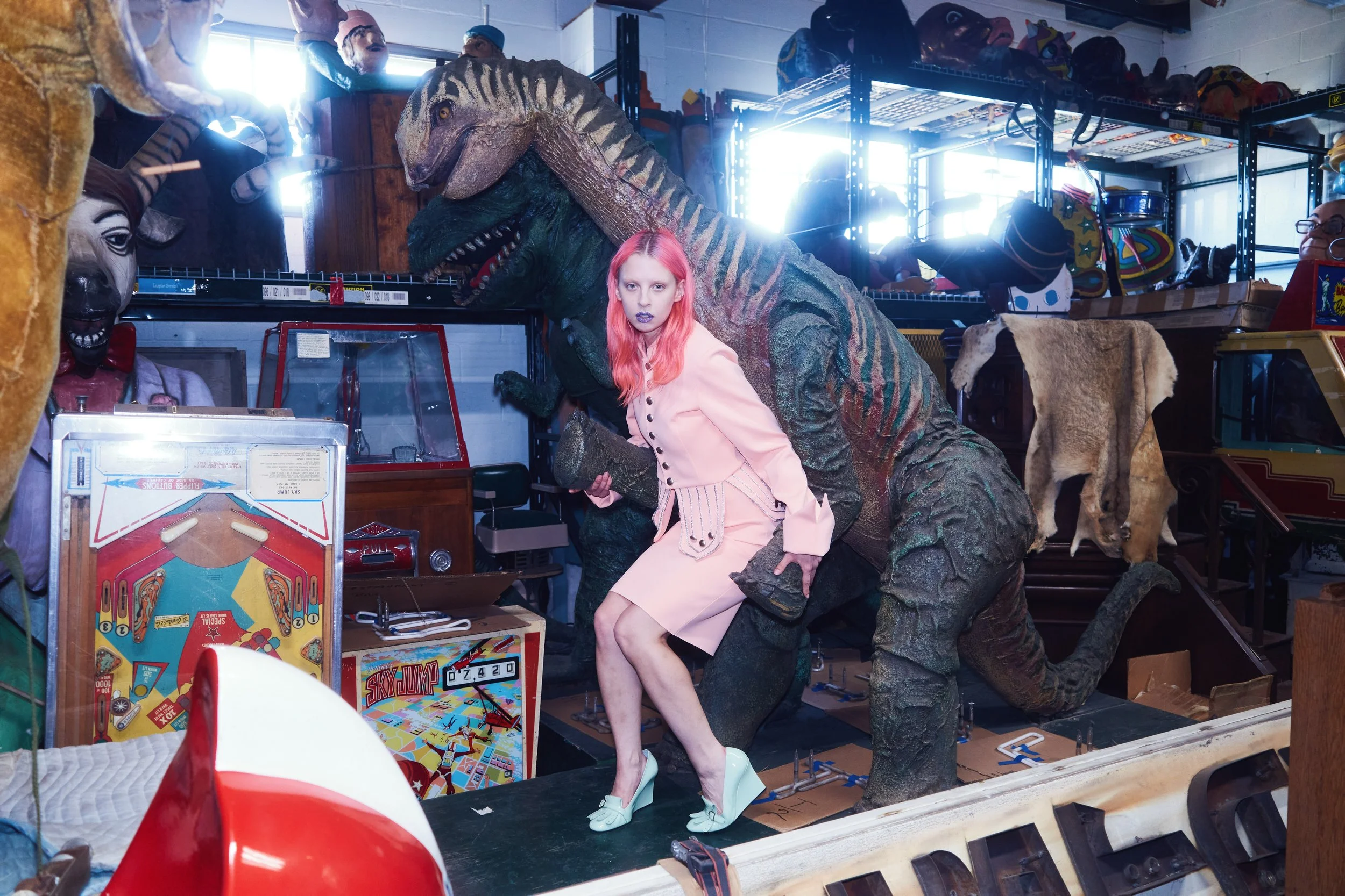 A woman with pink hair, wearing a light pink blazer and skirt, poses inside a storage room filled with vintage and toy items, including a large dinosaur figure, pinball machines, masks, and helmets.
