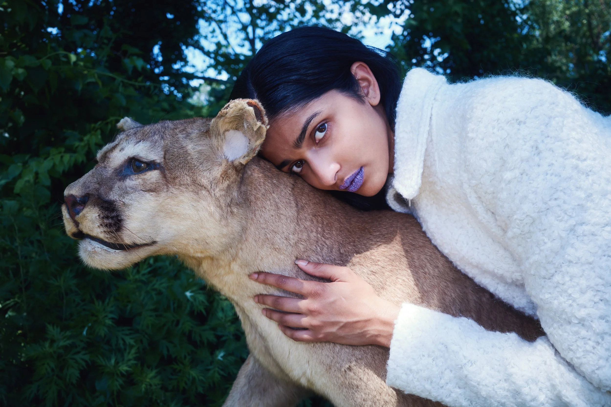 A woman with dark hair and purple lipstick cuddling a lion cub outdoors in a lush green environment.