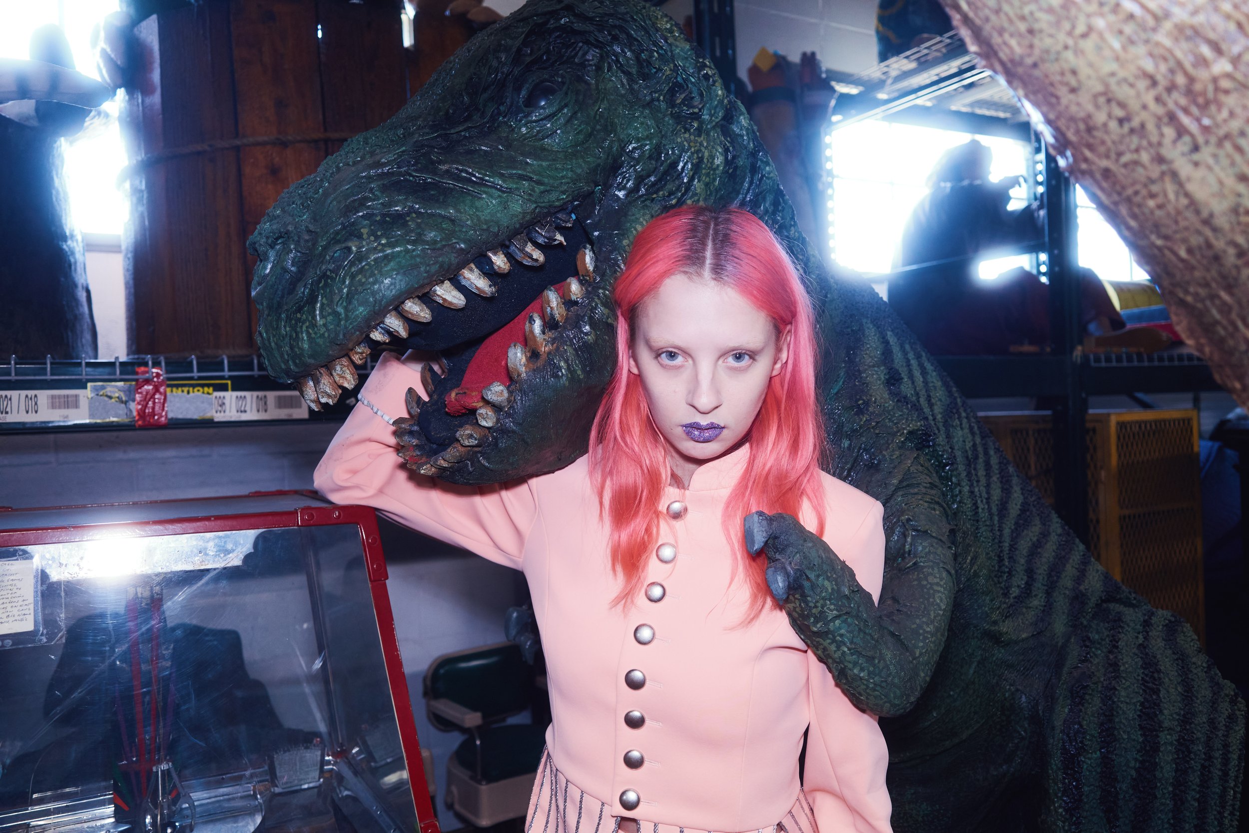 A woman with pink hair and purple lipstick posing with a life-sized model of a T-Rex dinosaur inside a room with wooden walls and storage shelves.