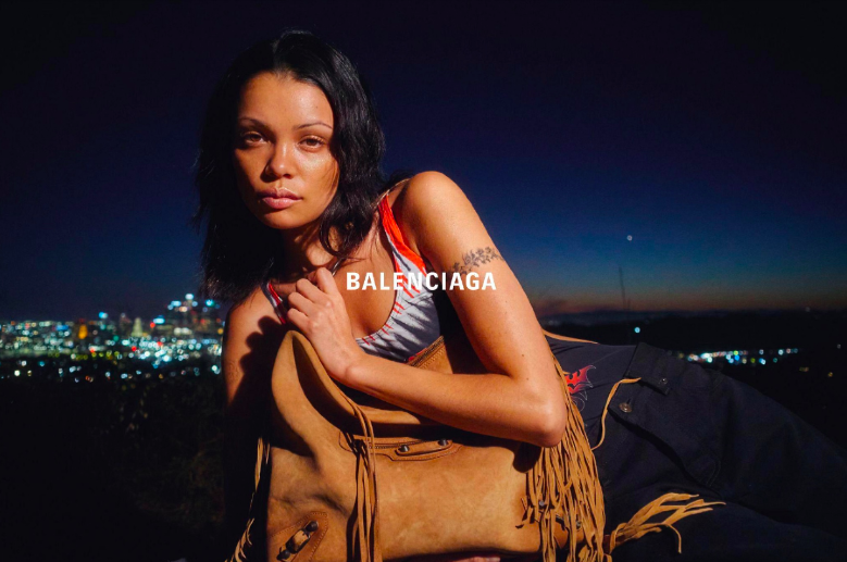 Woman with black hair, wearing a sleeveless top, sitting outdoors at night with city lights in the background, holding a brown fringed bag, and Balenciaga logo visible on her top.