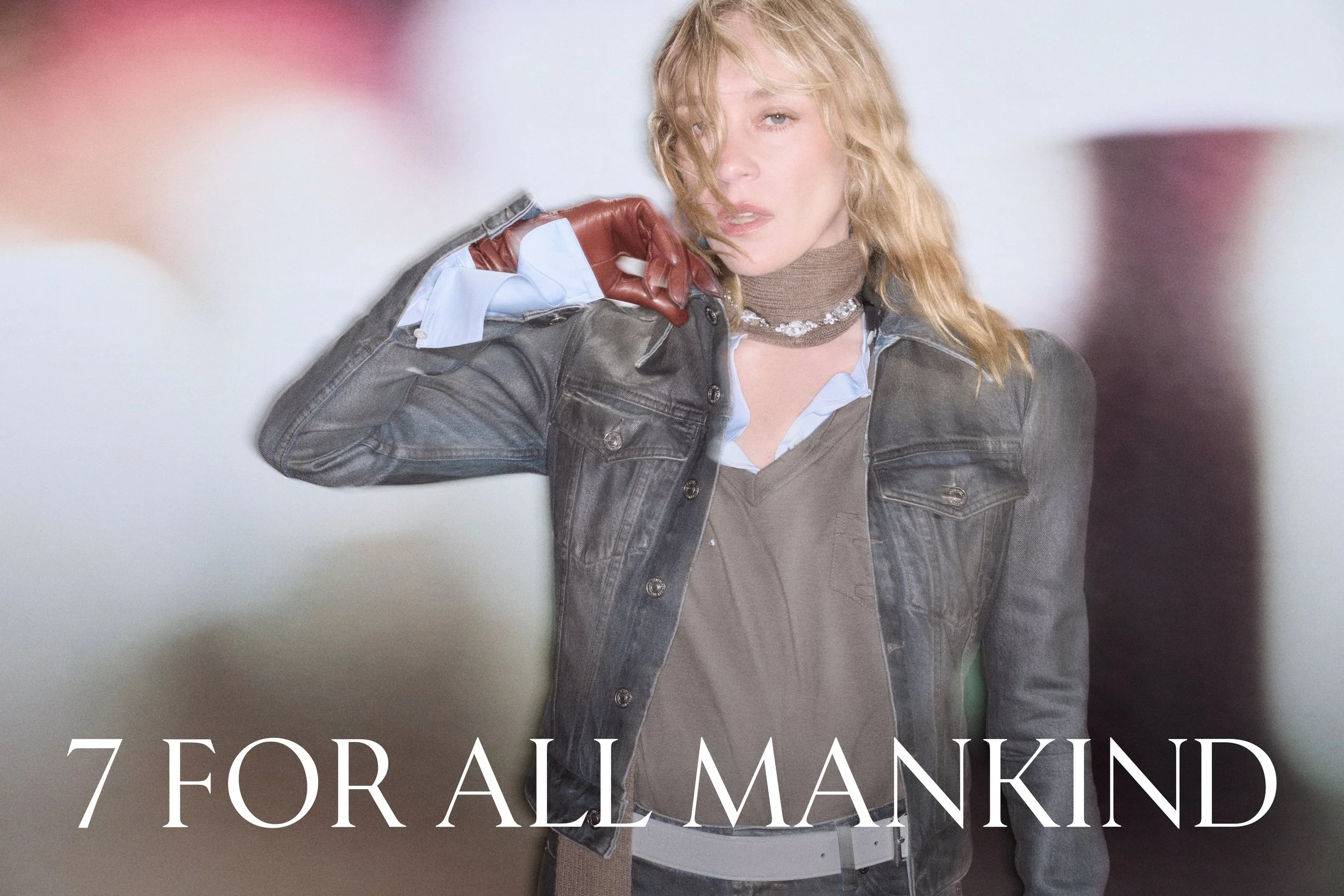 Close-up of a woman with curly blonde hair wearing a denim jacket, brown glove, and layered necklaces, looking into the camera with an intense expression.