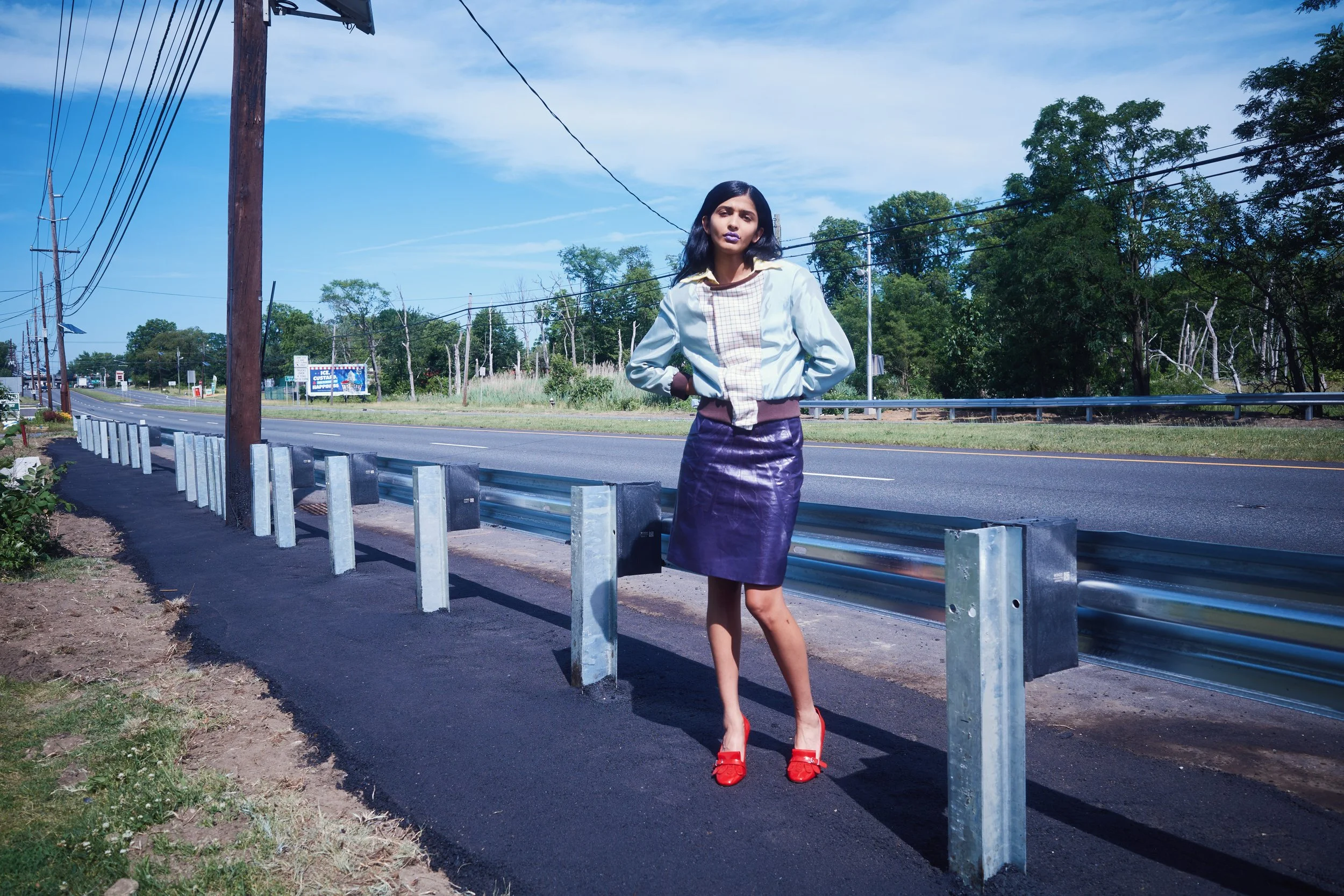 A woman standing on the side of a road between a metal guardrail and a grassy area. She has dark hair, is wearing a light-colored jacket, a shiny purple skirt, and red high heels. Behind her are trees and a blue sky with some clouds, along with utili