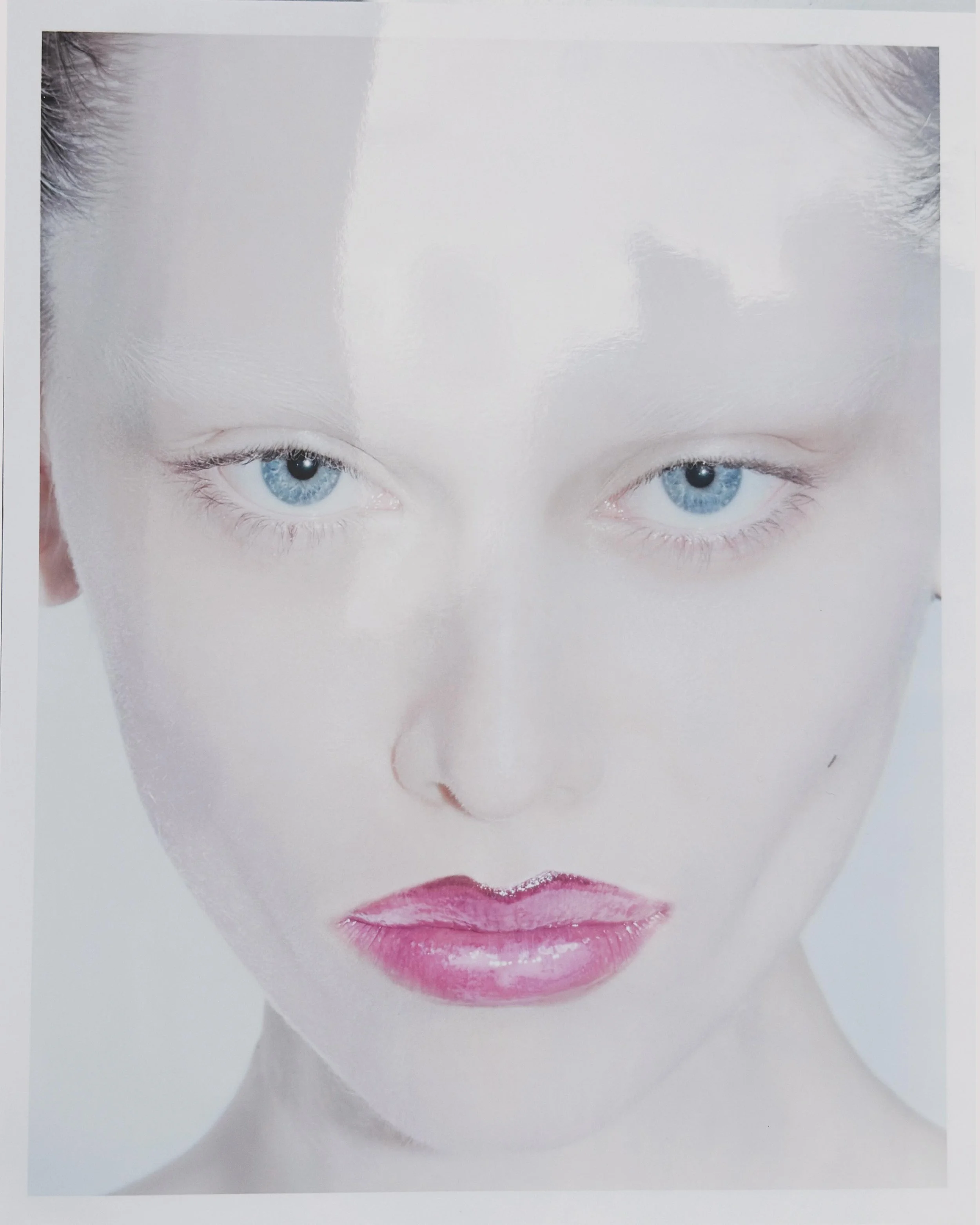 Close-up of a partially obscured woman’s face with blue eyes and pink lipstick, with shiny white hair and a blurred background.