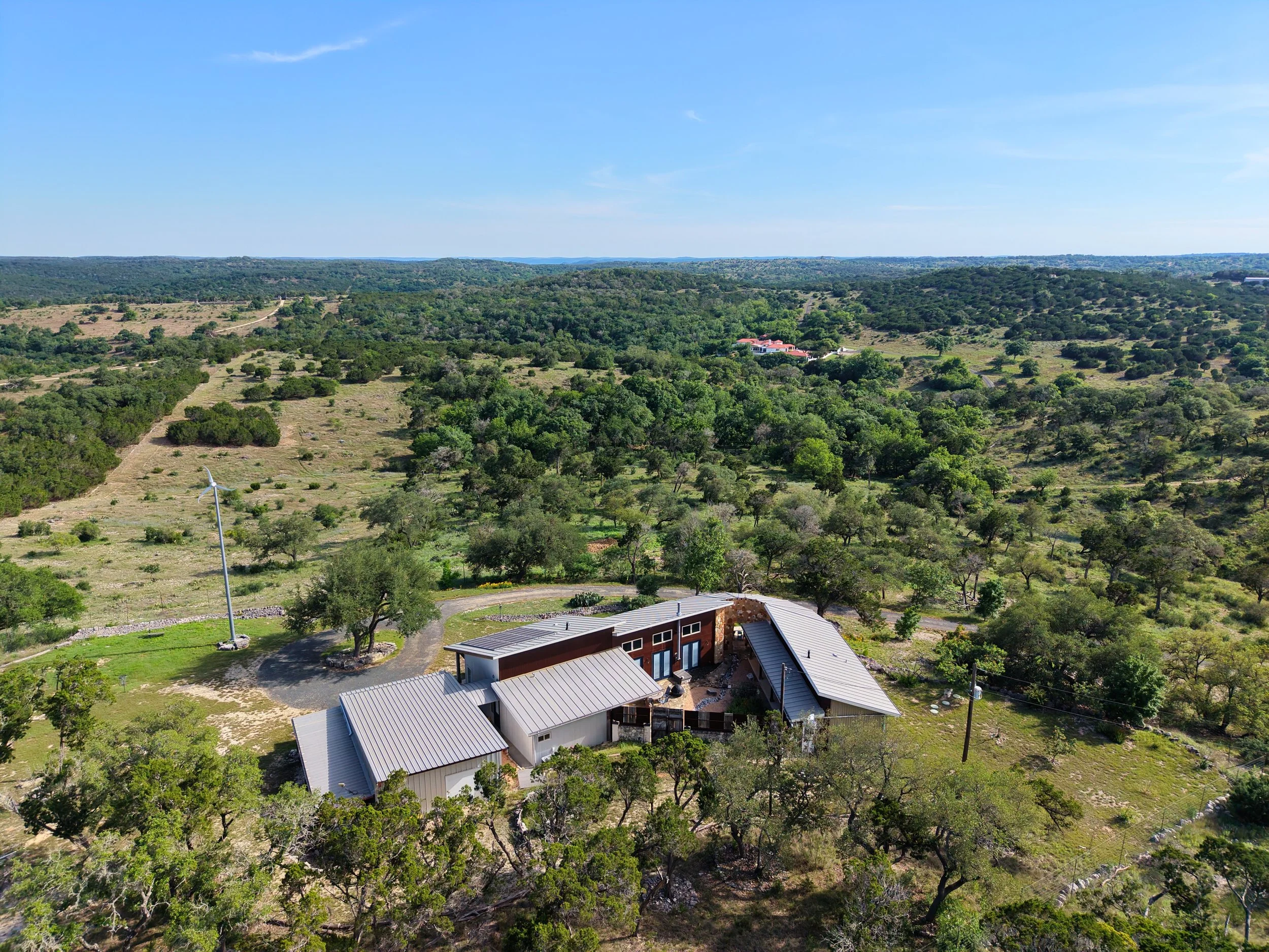 Aerial view of a rural landscape with hills, trees, a house, and wind turbines under a clear blue sky.