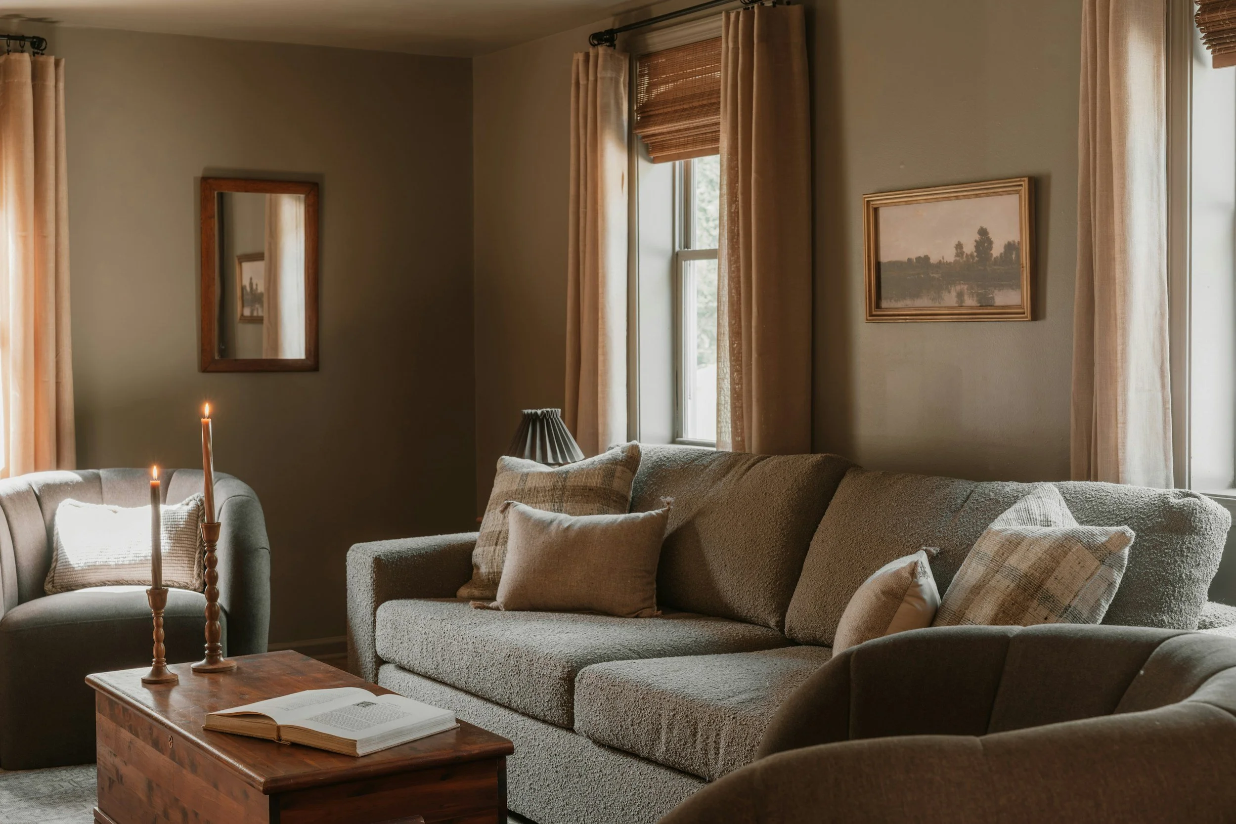 Living room with beige sofas, decorative pillows, wooden coffee table with candles and an open book, framed landscape paintings on wall, and windows with curtains and blinds letting in natural light.