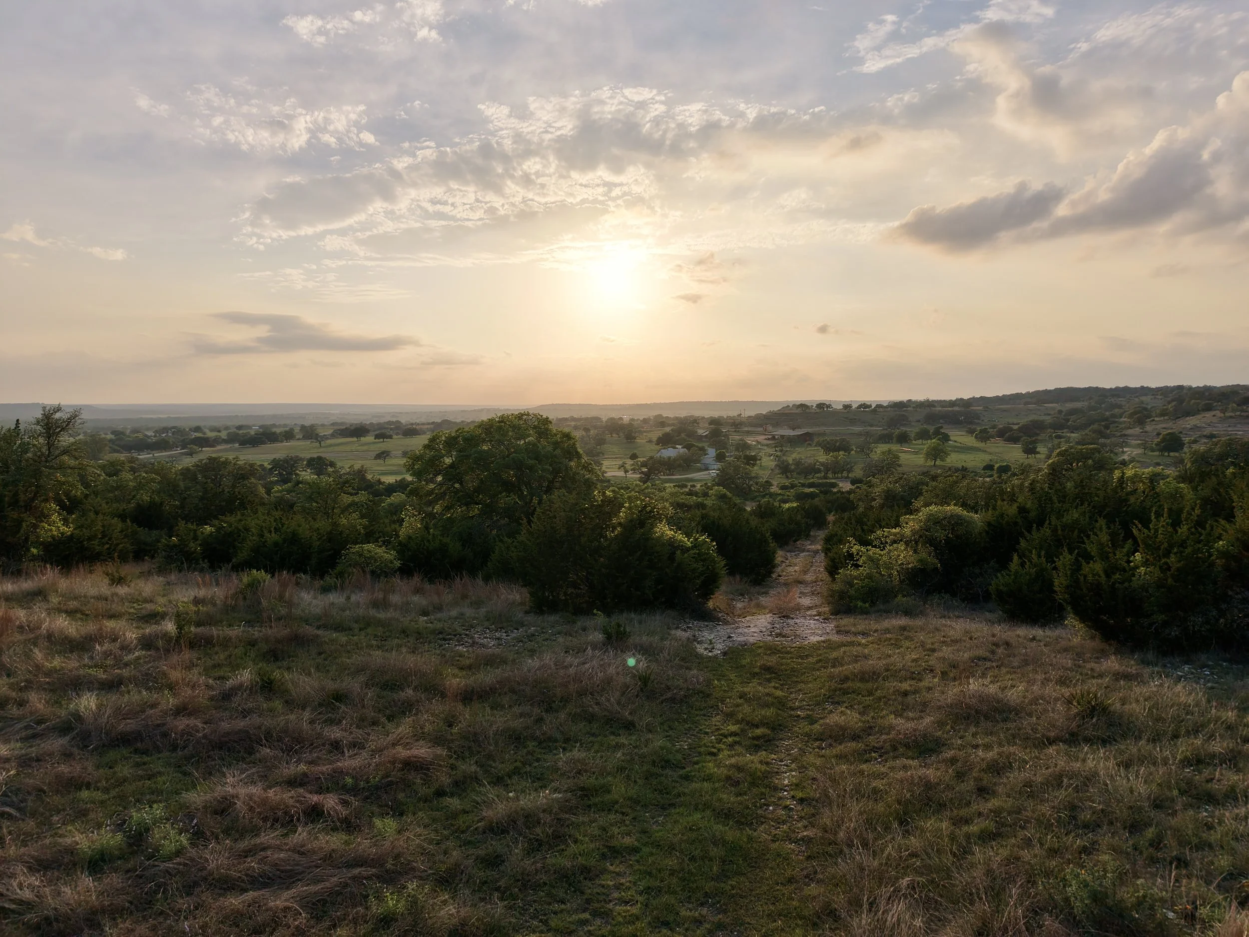 A scenic landscape during sunset showing rolling green hills, scattered trees, and a partly cloudy sky with the sun low on the horizon.