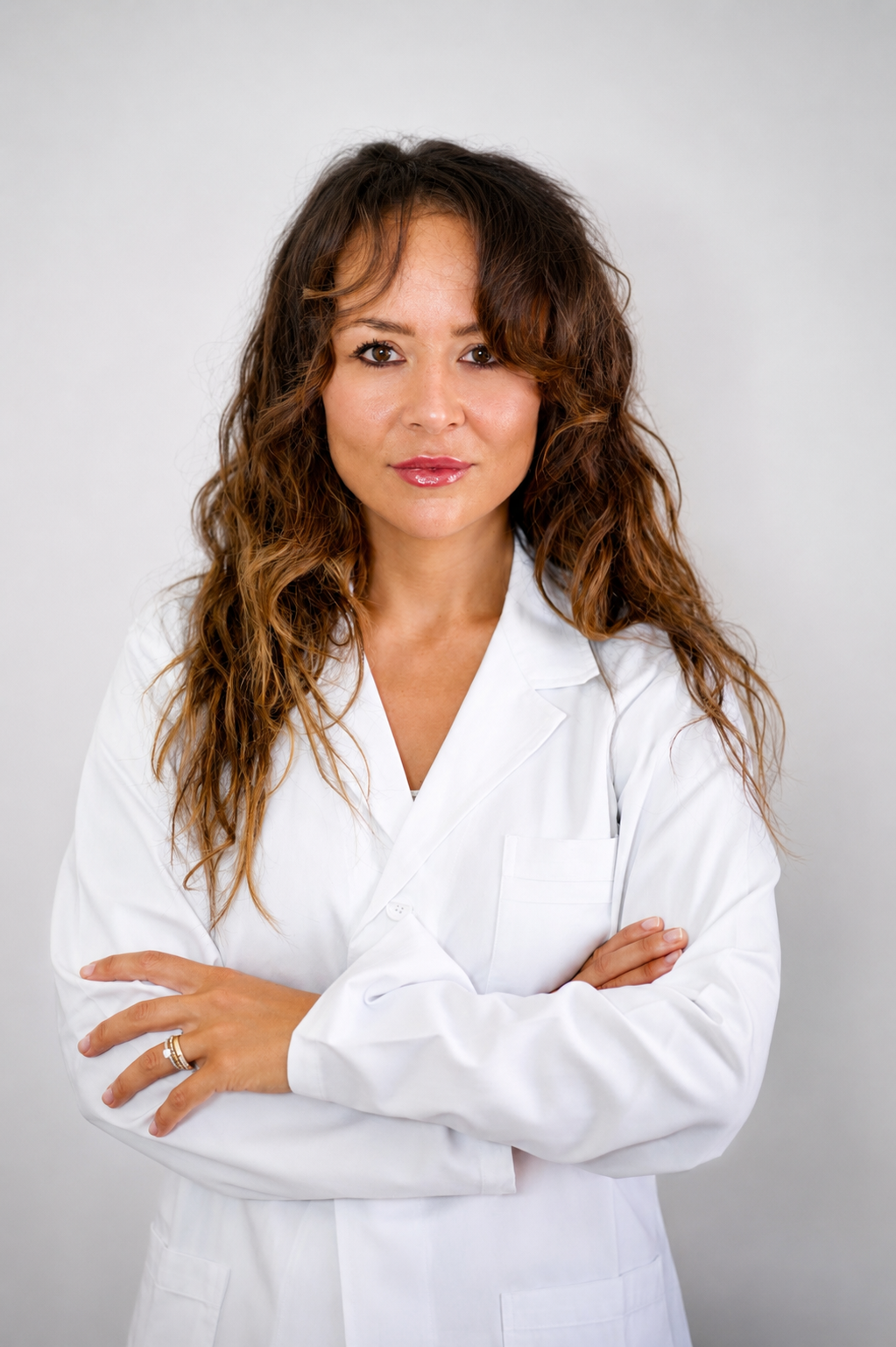 Donna con capelli riccioli scuri e occhi castani, vestita con un camice bianco, si trova contro uno sfondo neutro, con le braccia incrociate.