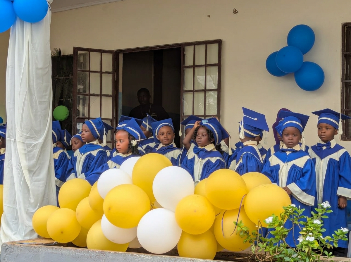 Children in blue and white graduation gowns and caps, standing behind yellow and white balloons, at a graduation ceremony.