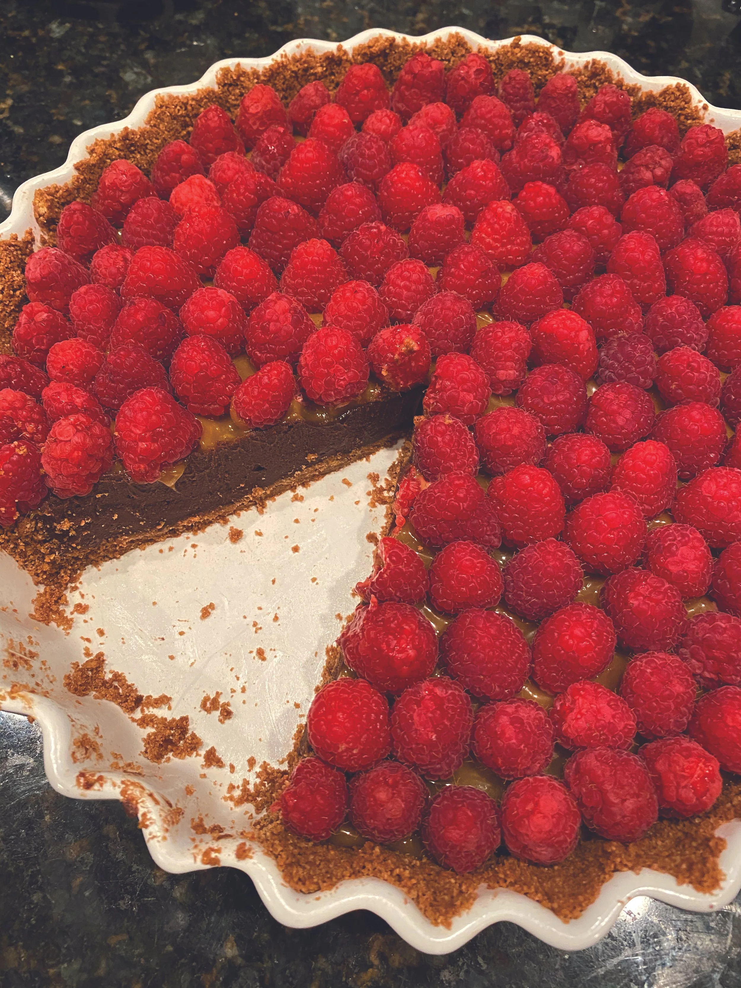 A pie with a chocolate crust, topped with fresh raspberries, on a white decorative dish on a dark countertop.