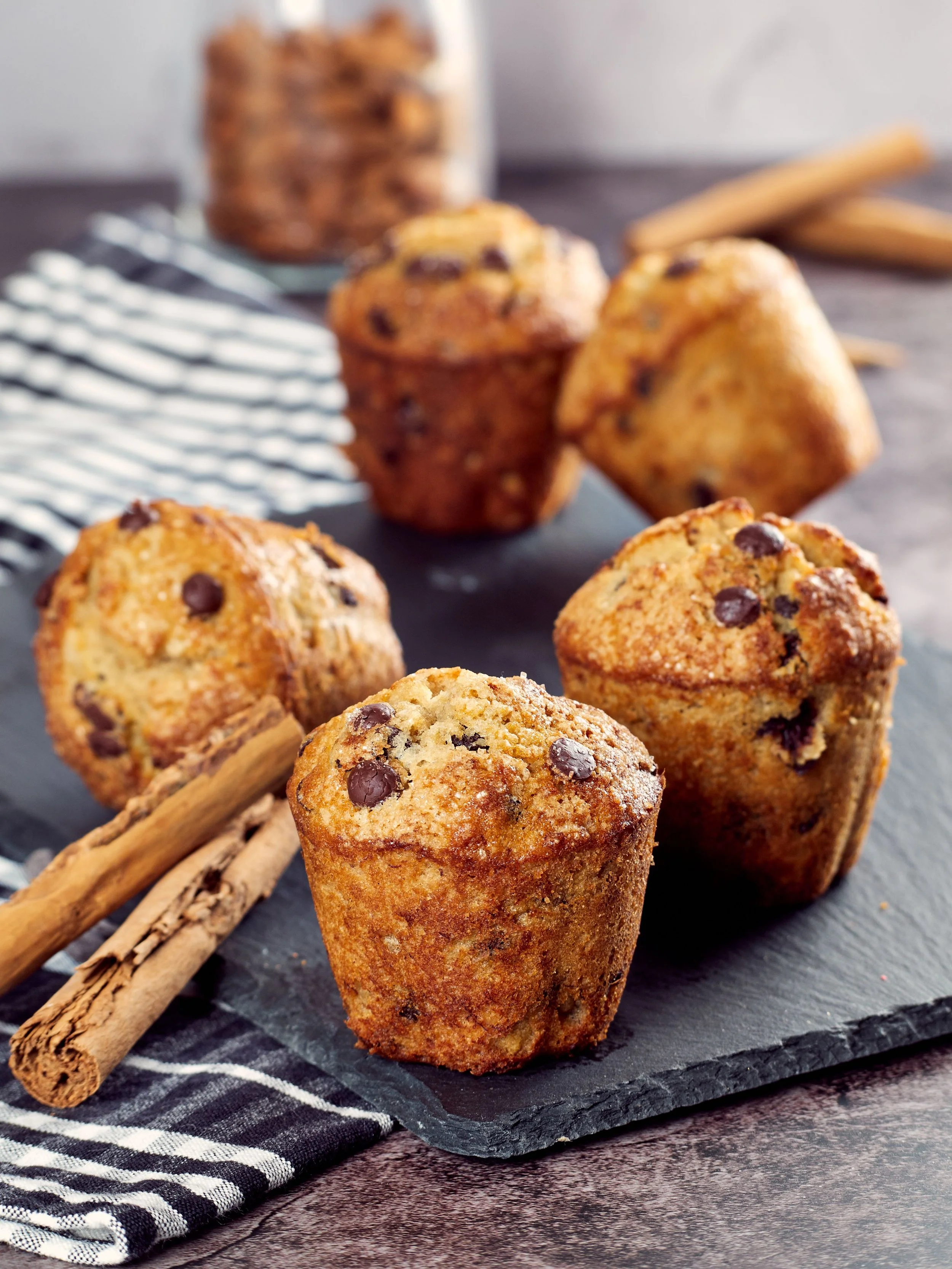 Chocolate chip muffins on a black slate serving board with cinnamon sticks and a black and white checkered napkin.