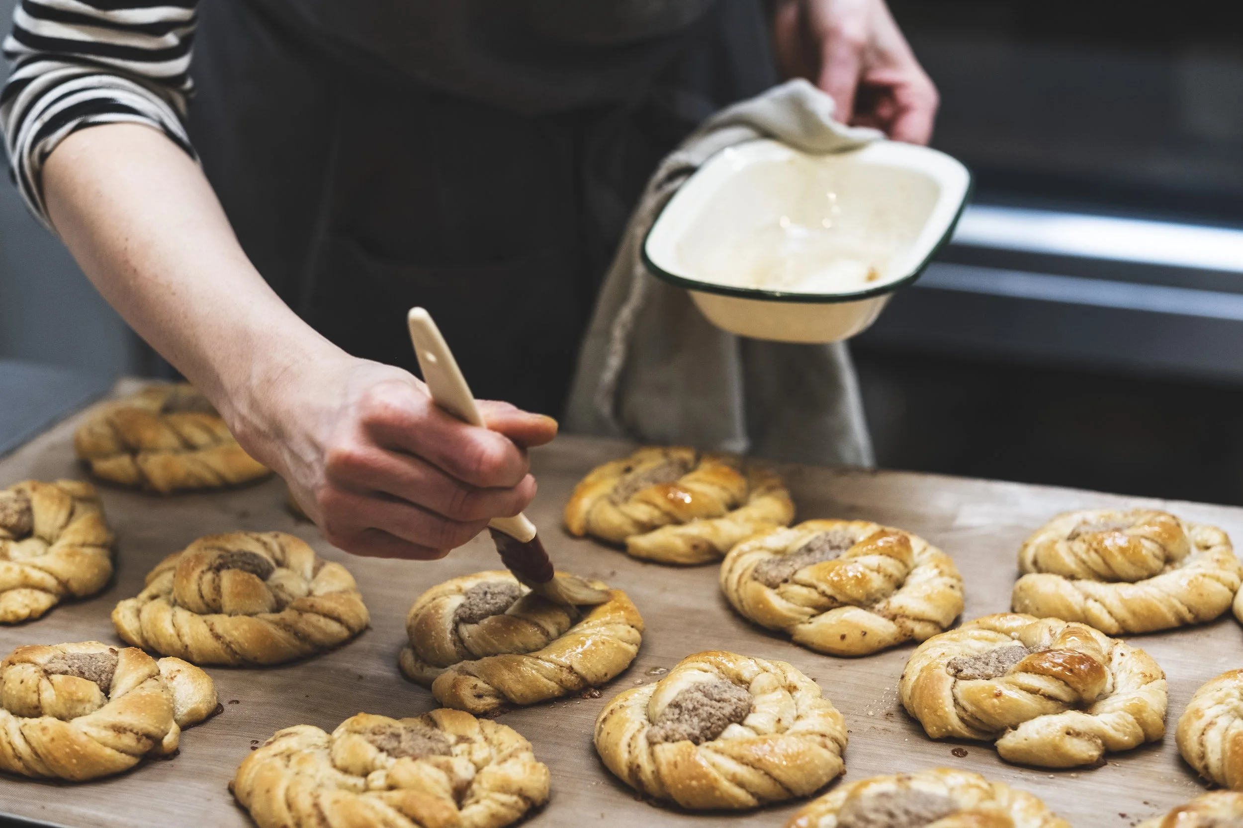 A person wearing a black apron and a striped shirt decorates freshly baked pastries with a squeeze bottle. The pastries are golden-brown with a twisted shape, filled with a dark filling, and topped with a glaze or powdered sugar. There are multiple pastries on a baking sheet or counter.