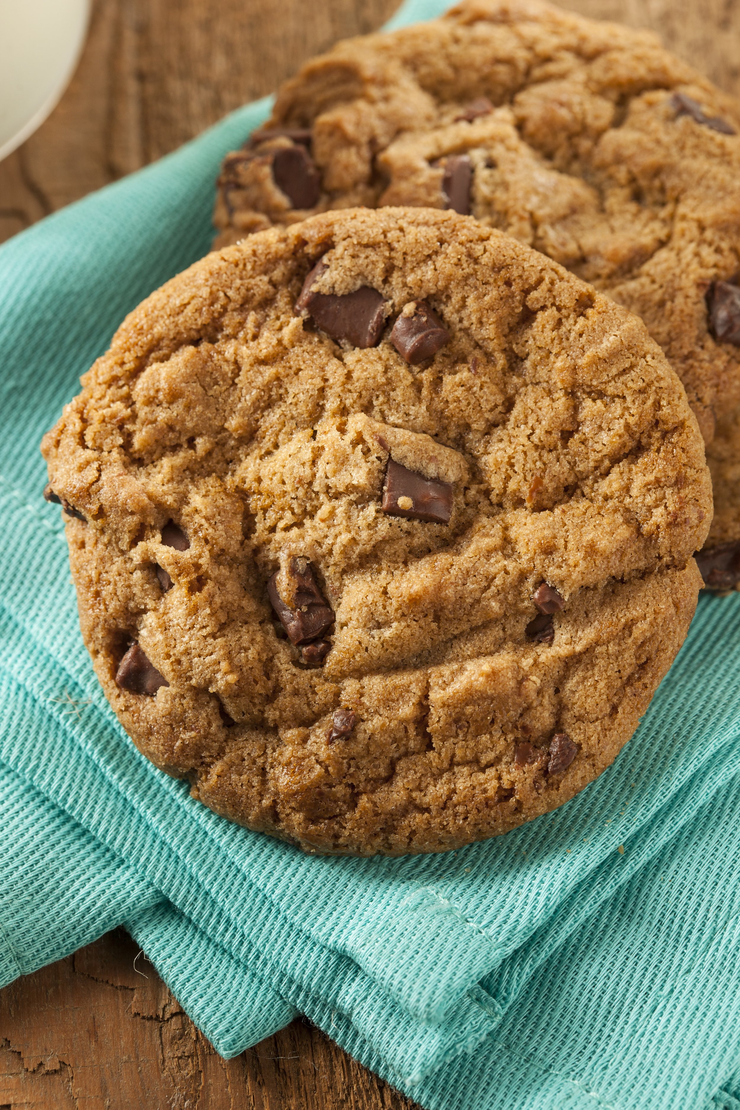 Two chocolate chip cookies on a teal cloth napkin placed on a wooden surface.
