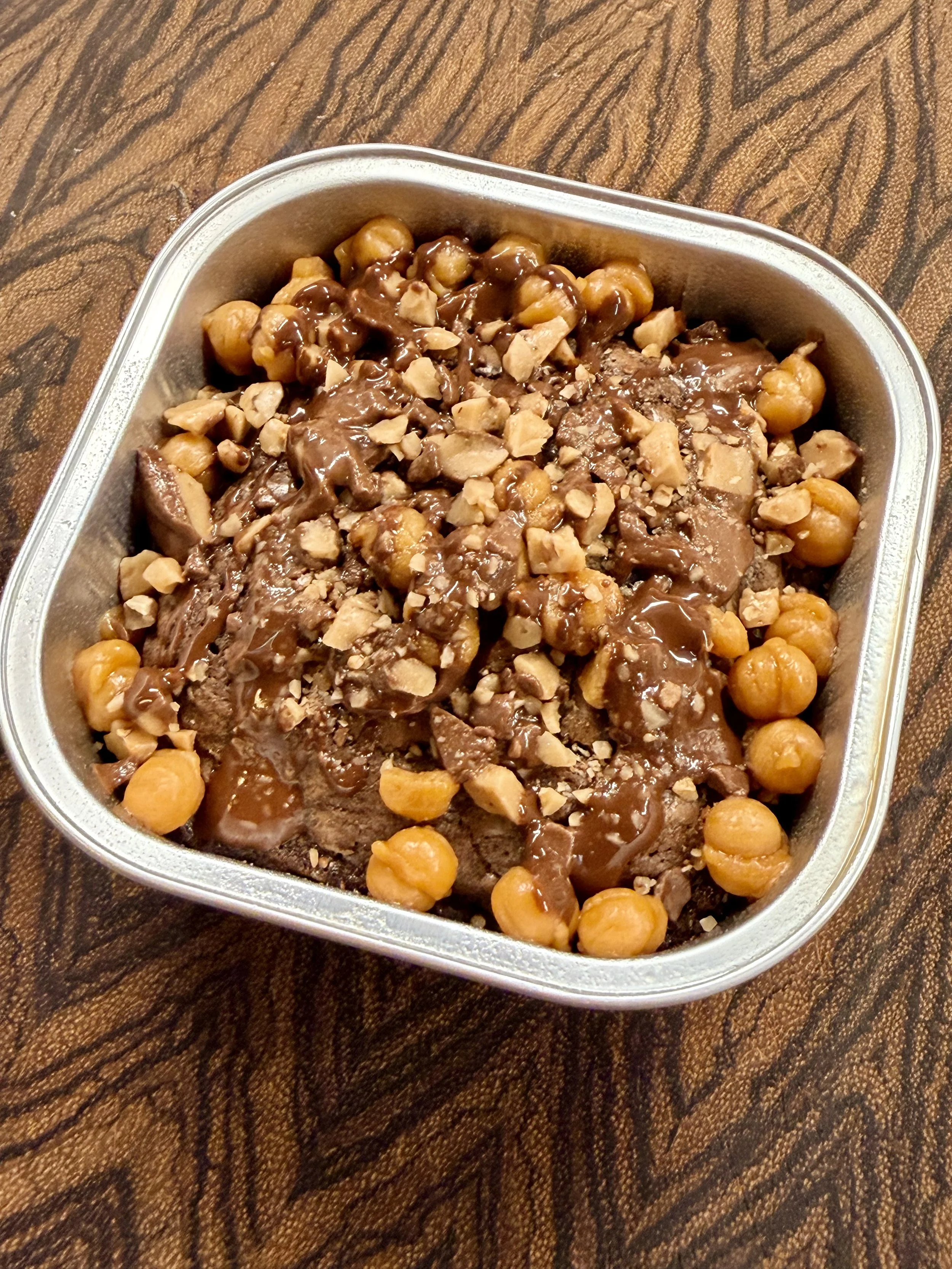 A metal container filled with chocolate dessert topped with chopped nuts and whole hazelnuts, on a wooden surface.