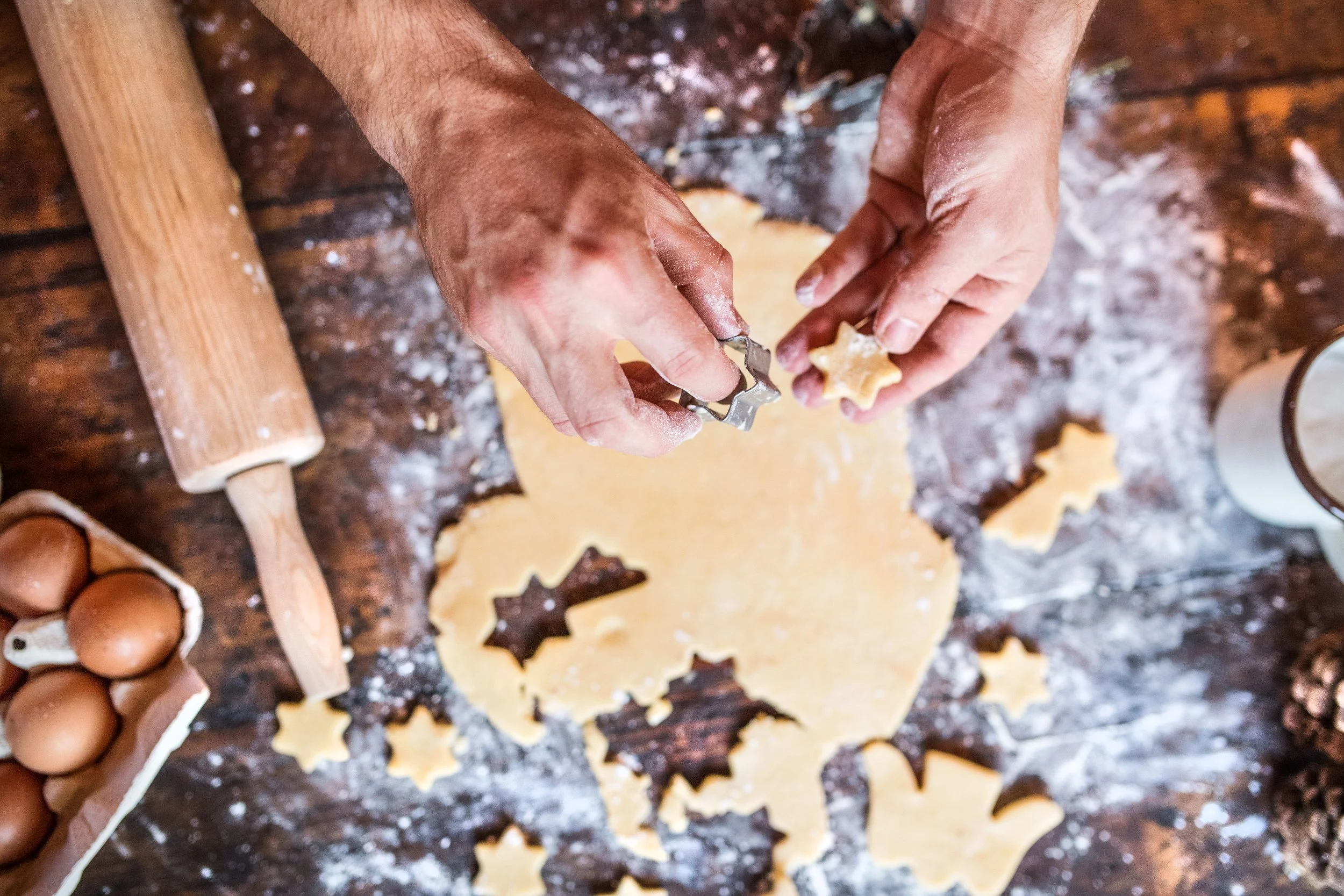 Hands pressing cookie cutter into rolled out cookie dough on a floured wooden surface, with a rolling pin nearby and eggs in the background.