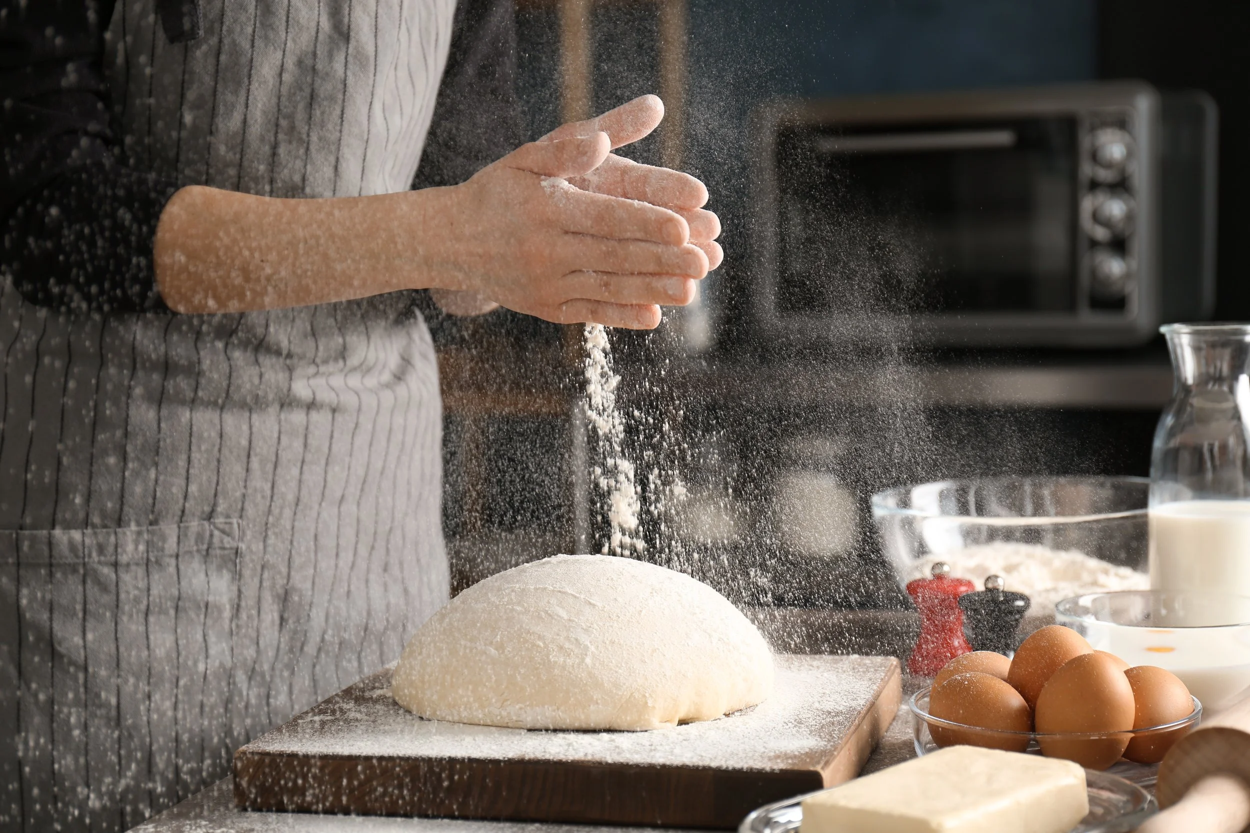 A person in a striped apron is clapping flour in a kitchen. There are baking ingredients on the counter, including a bowl of eggs, dough, and various baking supplies.