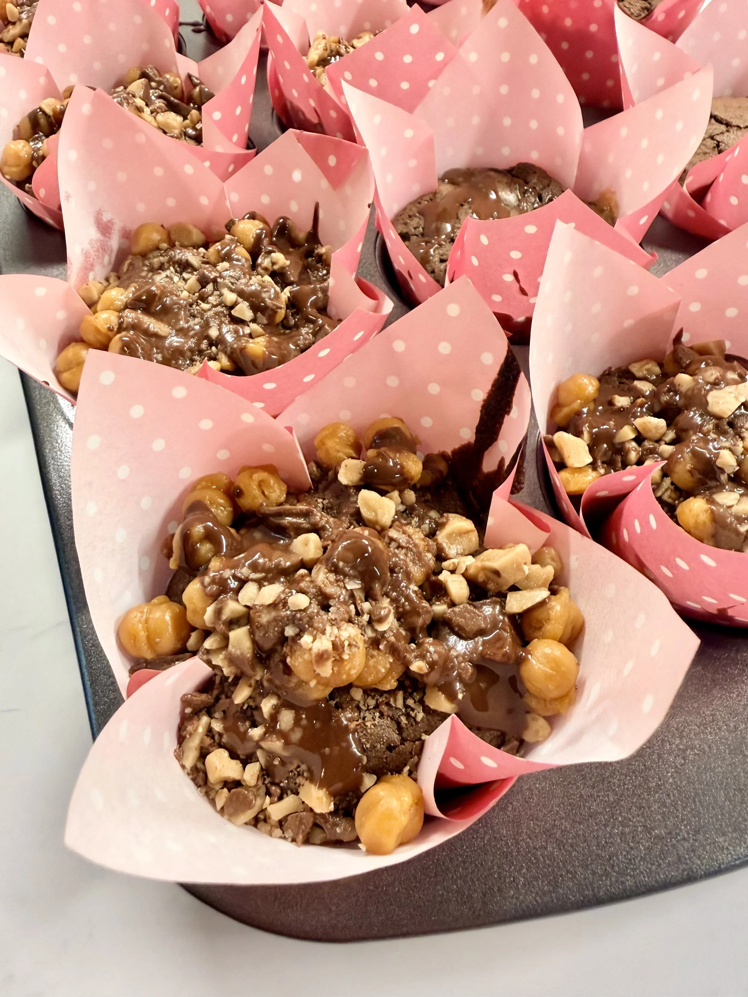 Assorted homemade muffins in pink polka dot paper liners decorated with chocolate, caramel, and chopped nuts.