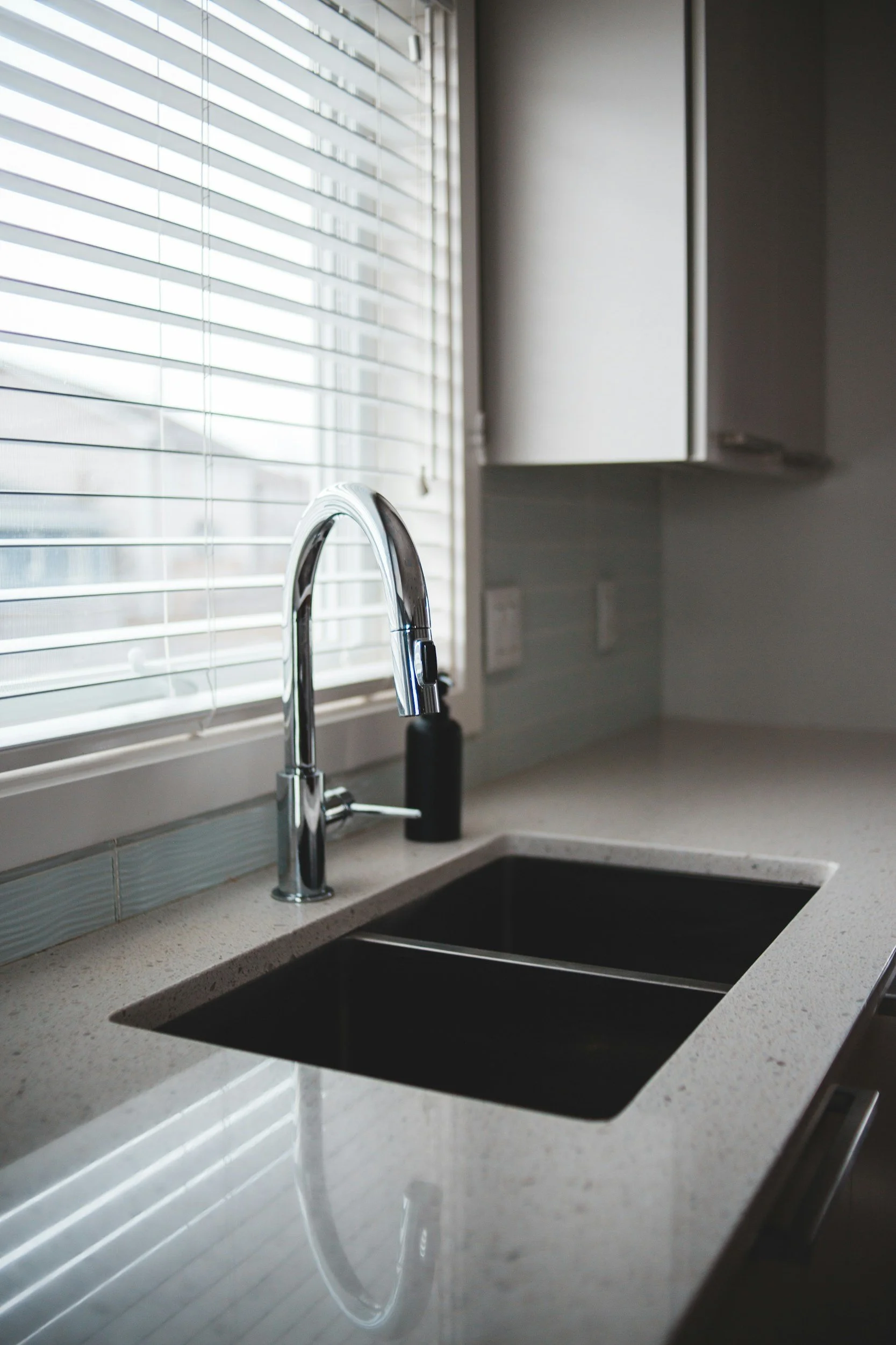 Close-up of a modern kitchen sink with a chrome faucet, set in a speckled beige countertop, and a window with white blinds in the background.