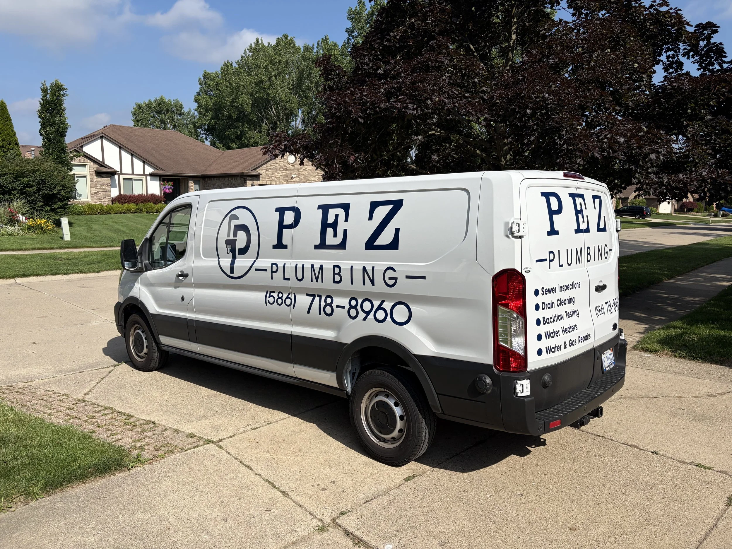 A white plumbing service van parked on a suburban street, with a house, trees, and a blue sky in the background.