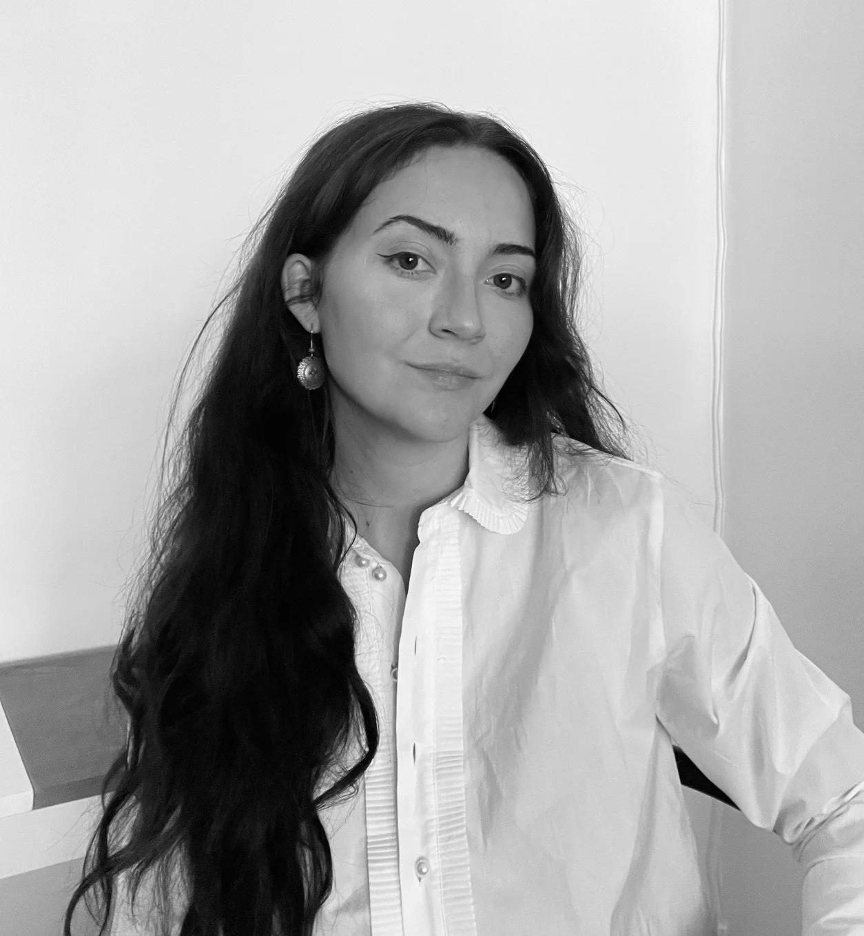 Black and white portrait of a woman with long wavy hair wearing a white shirt and statement earrings, sitting against a plain wall.