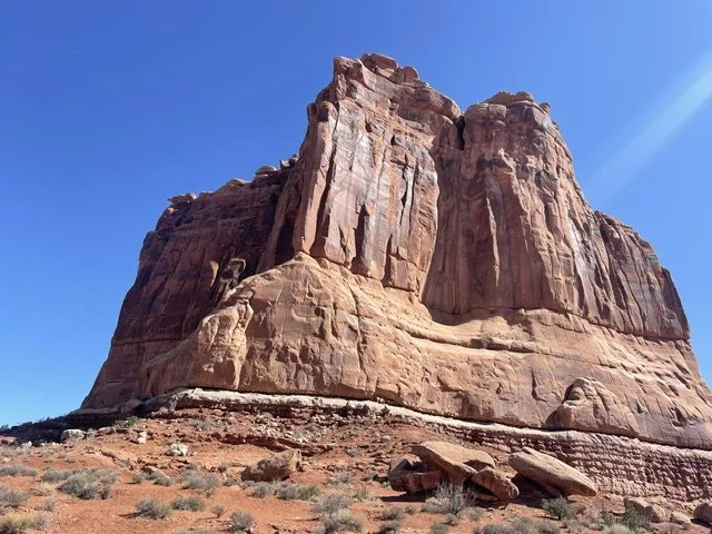 A large red rock formation in a desert landscape with sparse vegetation under a clear blue sky.