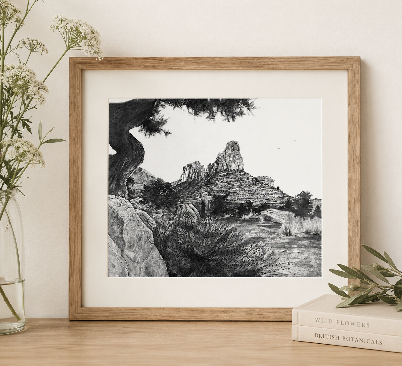 Black and white landscape illustration of rocky terrain with mountain peaks, trees, and a large gnarled tree in the foreground, framed in a wooden frame.