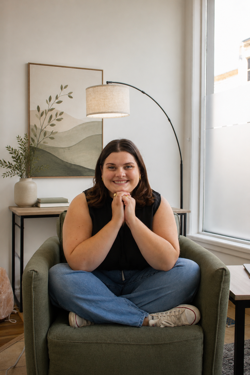 Smile woman sitting cross-legged on a green armchair with hands clasped under chin in a well-lit room with a window, framed artwork, a potted plant, and a floor lamp.