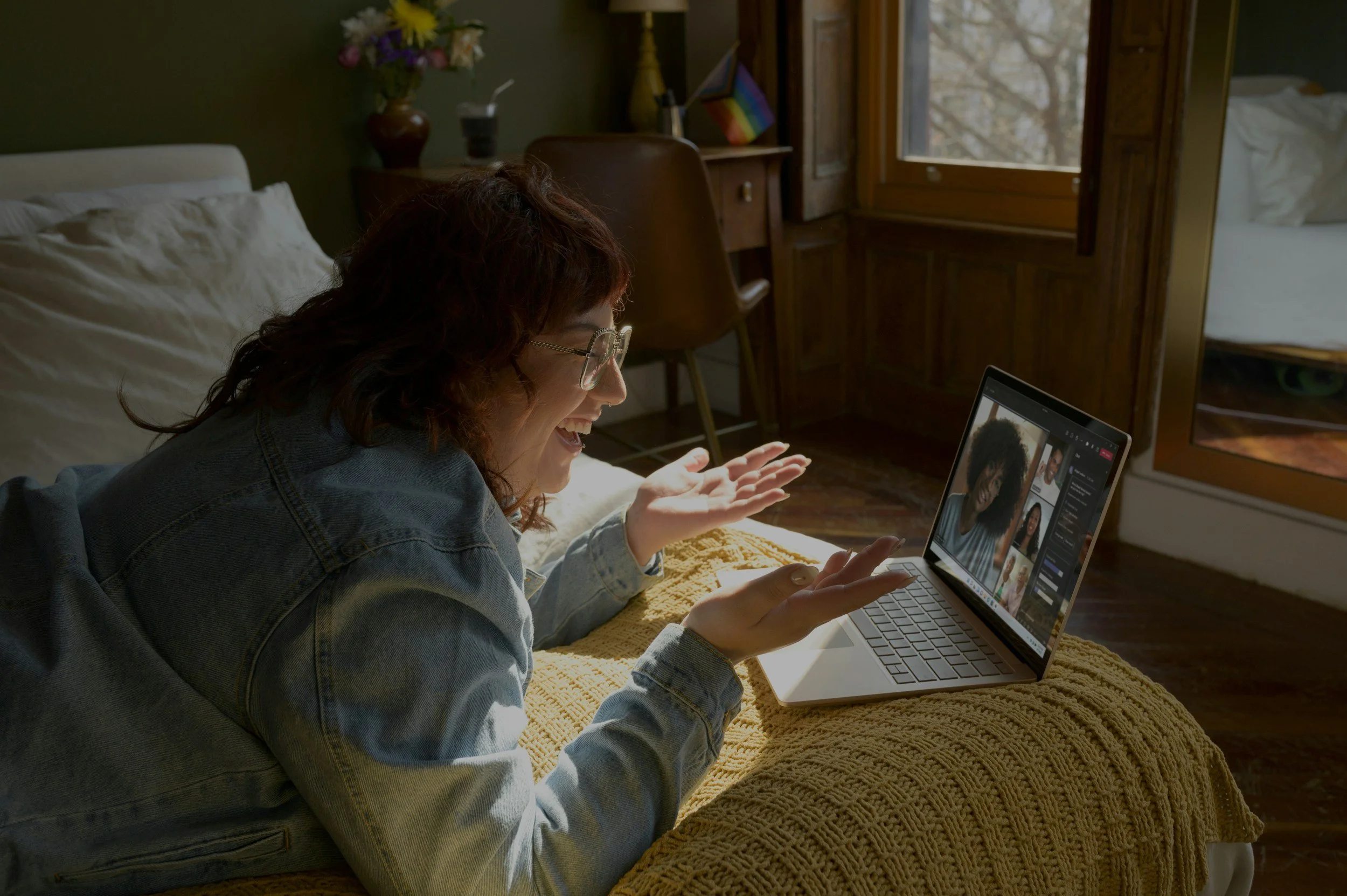 A woman with dark, curly hair and glasses smiling and engaging in a video call on her laptop while lying on her side on a bed covered with a yellow blanket. The room has wooden furniture, a window, and some decorative items including a vase with flowers.
