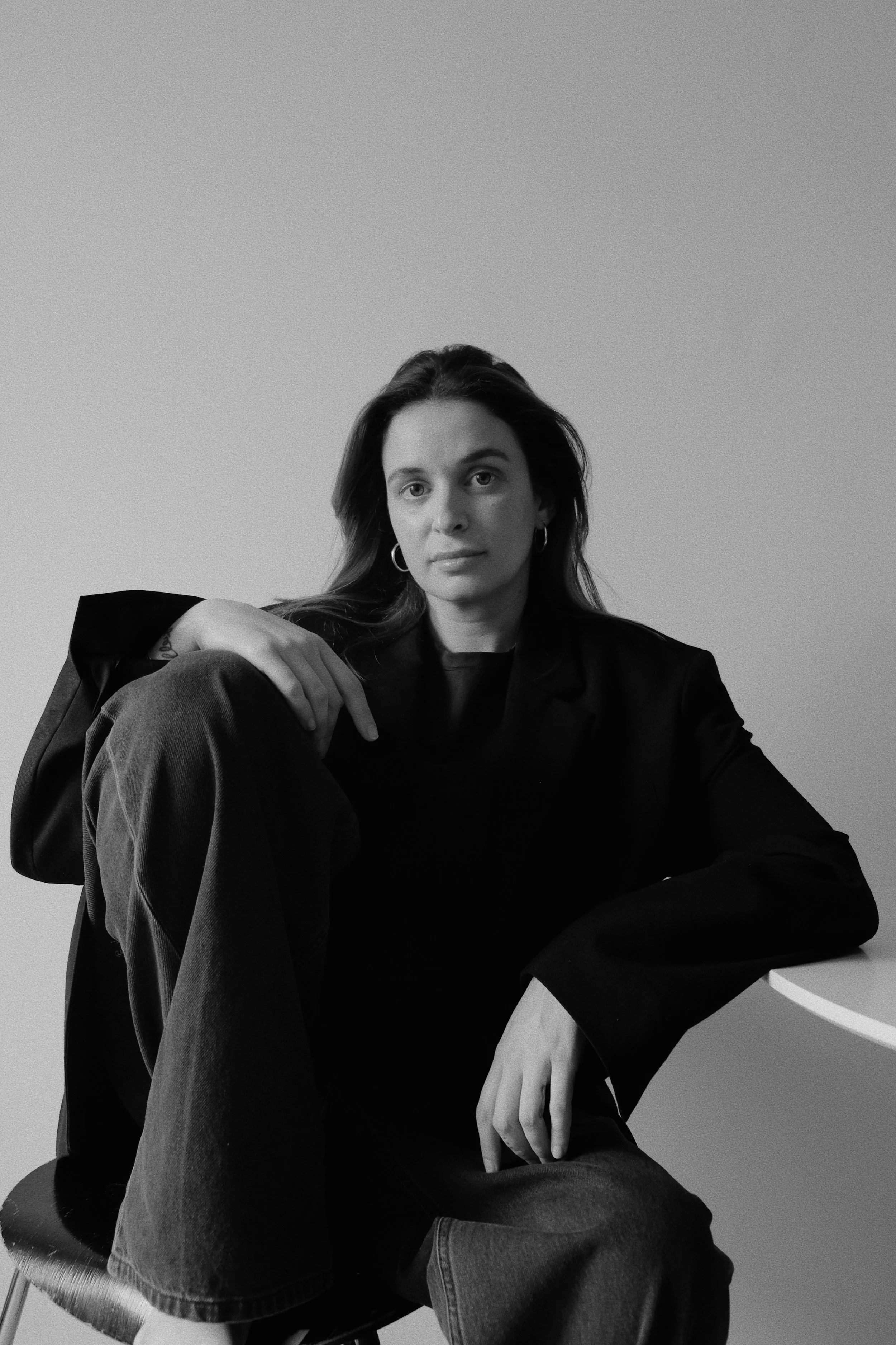 Schwarz-Weiß-Foto einer jungen Frau, die entspannt auf einem Stuhl sitzt, mit einem lockeren schwarzen Blazer und Jeans, in einem minimalistischen Raum mit hellem Hintergrund.