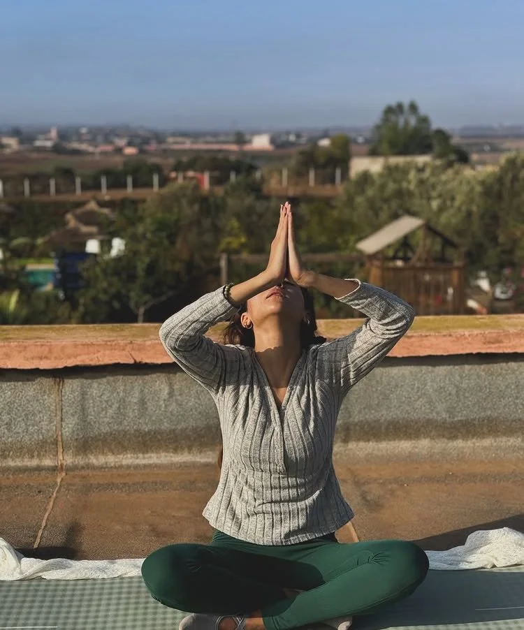Une femme pratique la méditation ou la prière en position assise en plein air, avec les mains jointes au-dessus de la tête, sur un toit ou une terrasse, avec un paysage de campagne en arrière-plan.