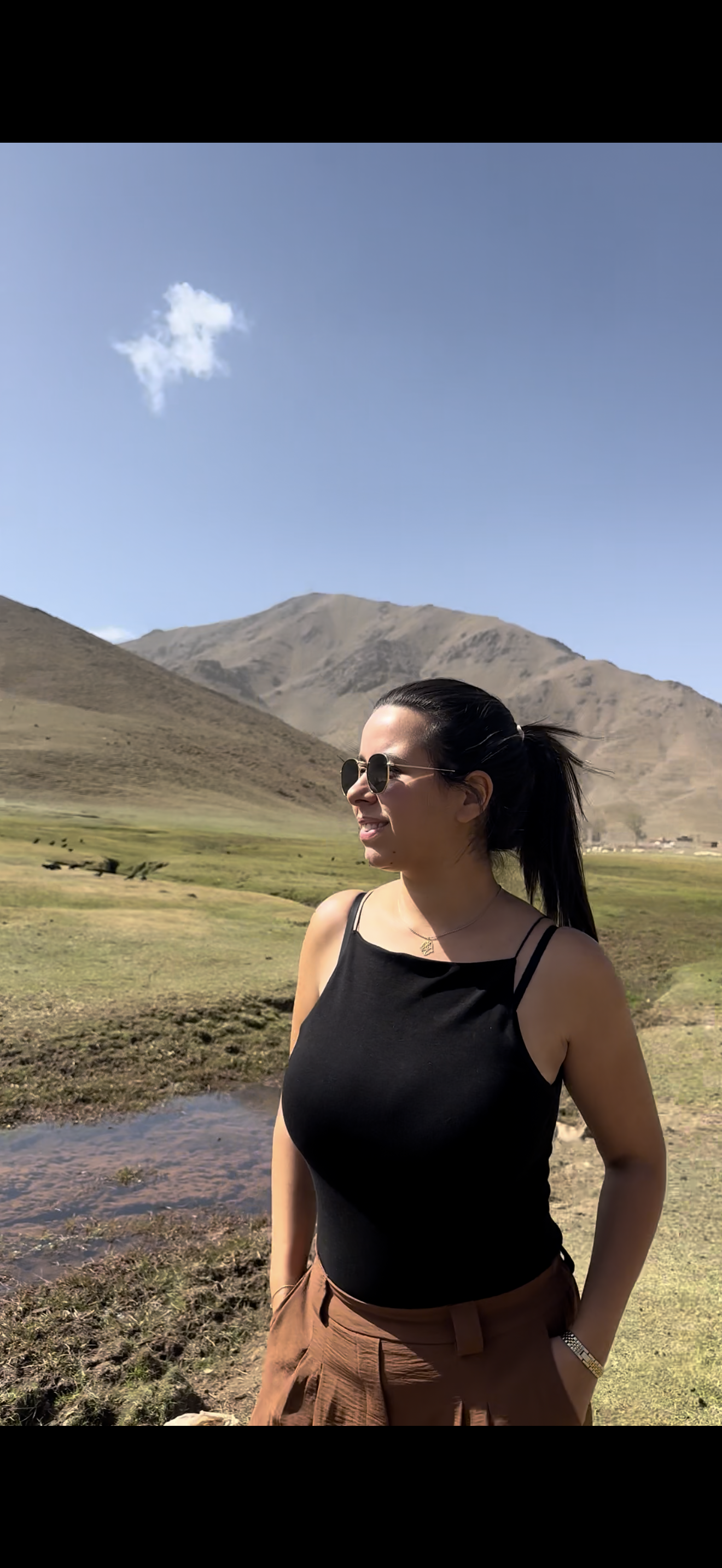 Femme souriante portant des lunettes de soleil et un haut noir, debout dans un paysage de montagnes, herbe et ciel bleu avec peu de nuages.