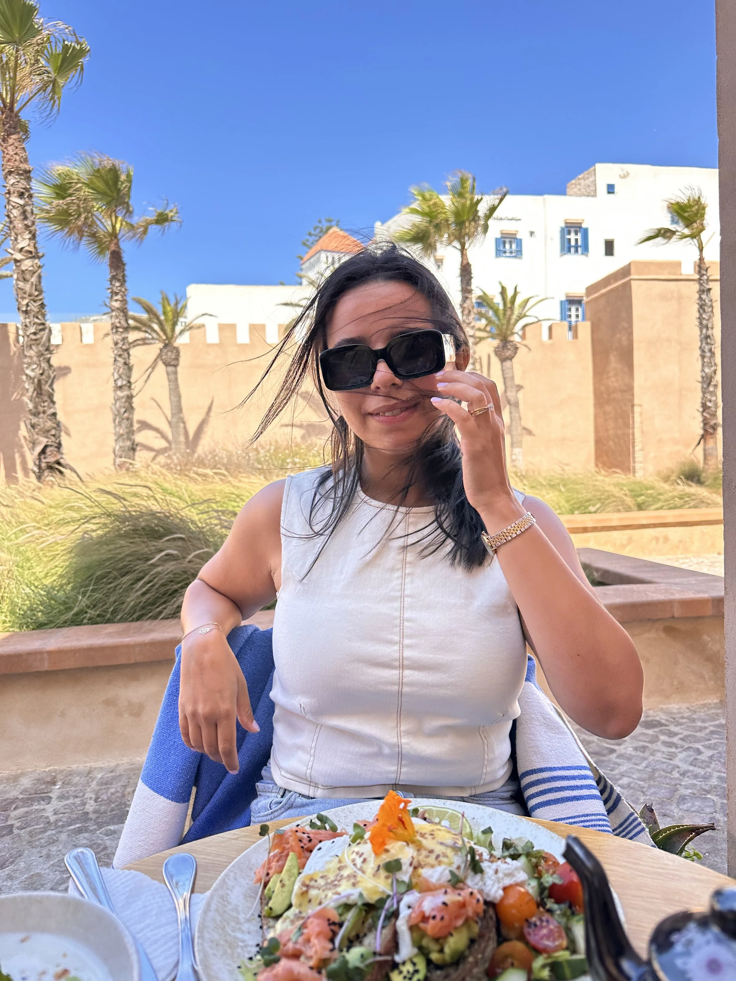 Une femme portant des lunettes de soleil, assise à une table en extérieur, avec un plat de nourriture fraîchement préparée devant elle, dans un cadre ensoleillé avec des palmiers et des bâtiments en arrière-plan.
