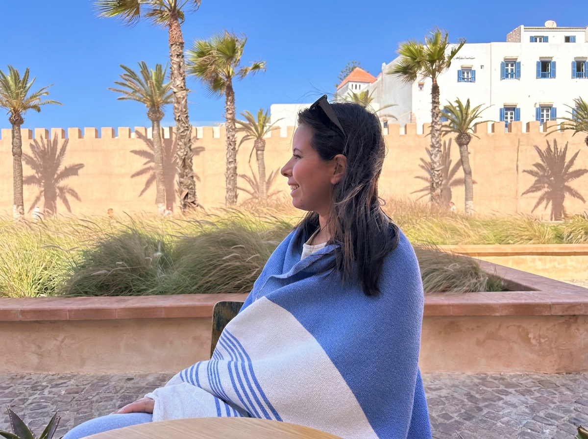 Femme souriante assise en plein air, enveloppée dans une serviette bleue et blanche, avec un décor de palmiers, mur beige, et bâtiments blancs en arrière-plan, sous un ciel bleu.