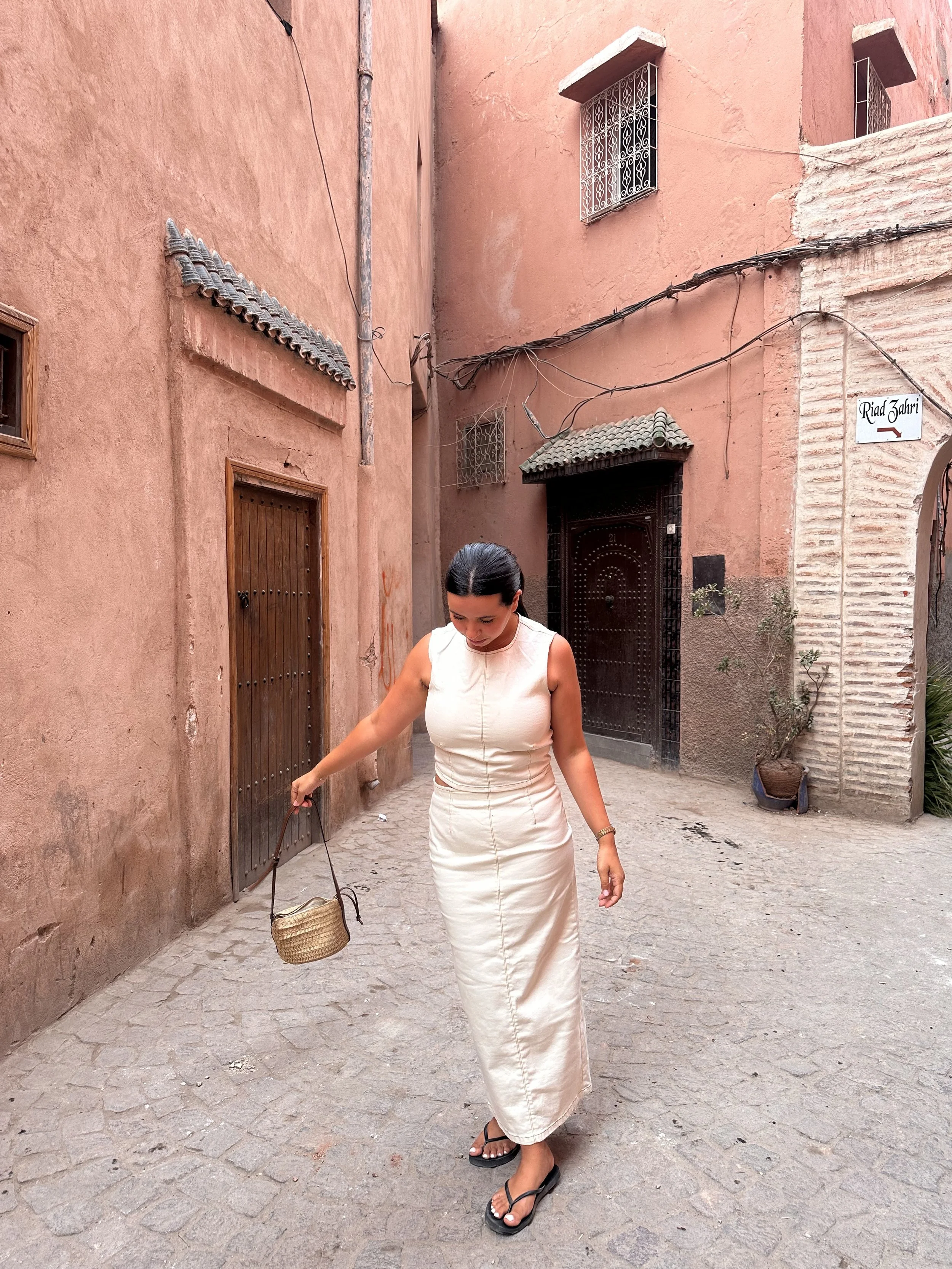 Une femme marchant dans une ruelle pavée avec des murs en terre cuite, portant une robe longue beige, des sandales à talons et tenant un sac en paille, devant une porte en bois et une fenêtre avec des barres en métal.