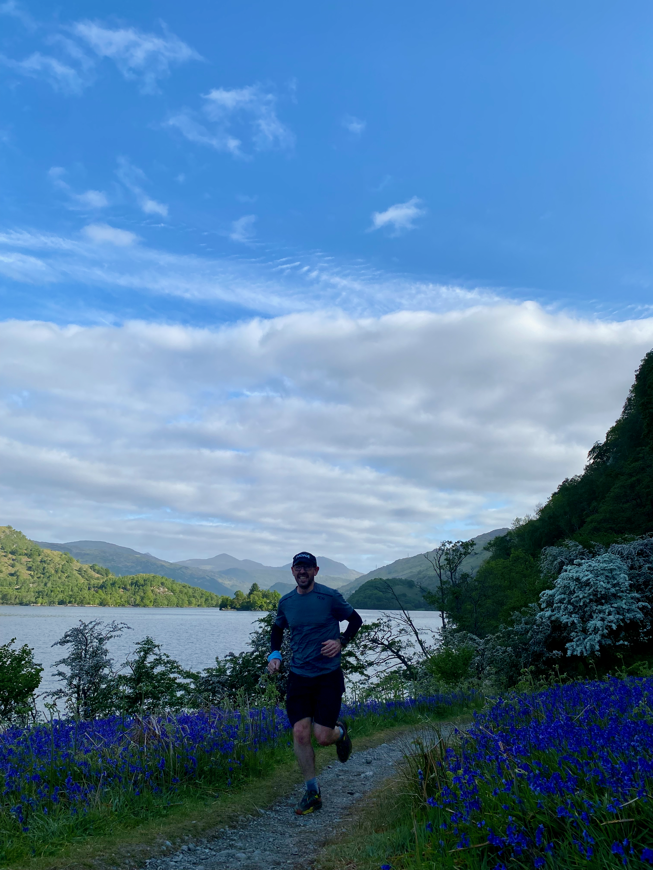 A man running on a trail beside a lake with mountains and trees under a partly cloudy sky.
