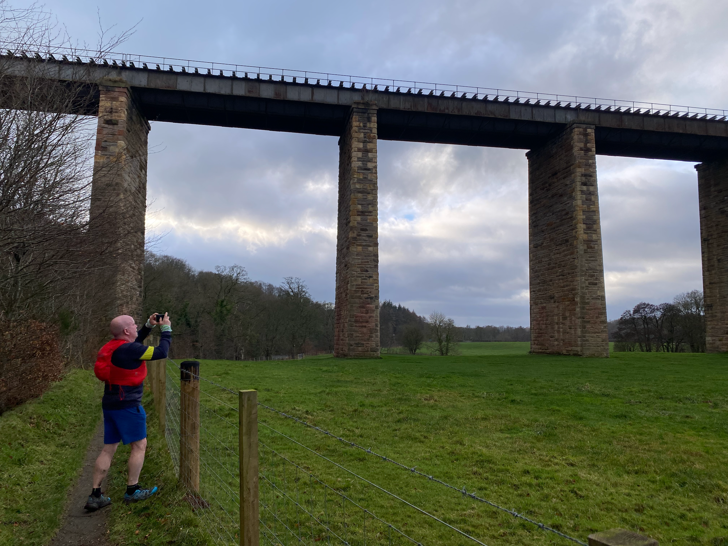 A man in a red and blue jacket taking a photo with his phone while standing on a trail next to a wire fence, overlooking a field with a large stone bridge with tall piers and a railway track on top, against a cloudy sky.