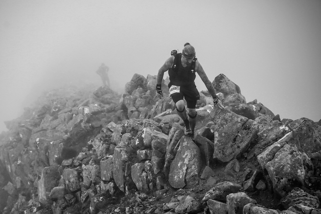 A mountain climber in a black tank top and shorts climbing over large rocks, with a foggy background and another climber visible in the distance.