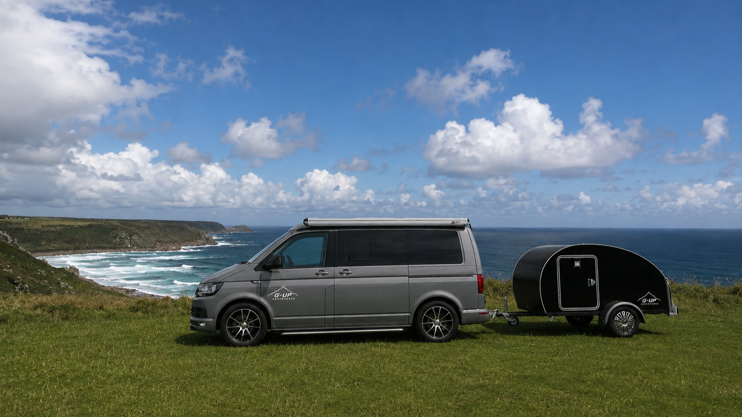 A gray van towing a black small trailer with a door, parked on a grassy area overlooking the ocean with cliffs and a partly cloudy sky.