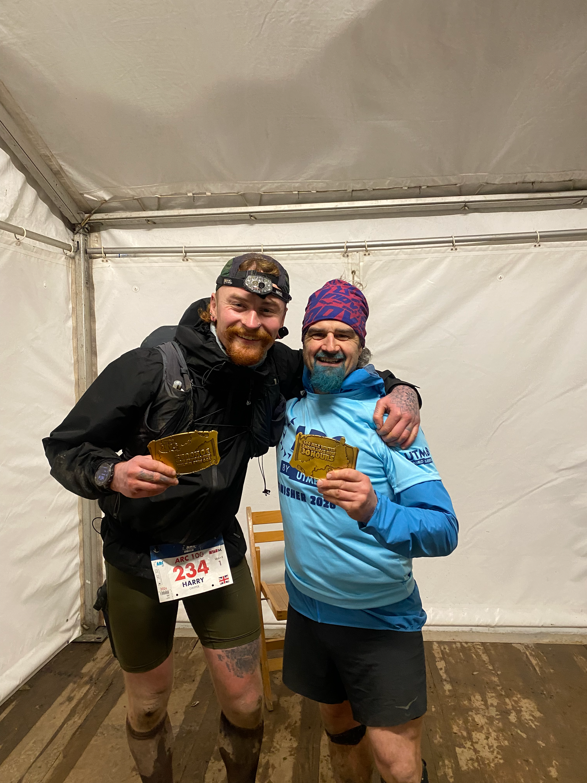 Two men smiling and celebrating, holding medals at a race event inside a tent, both wearing running gear and race bibs.