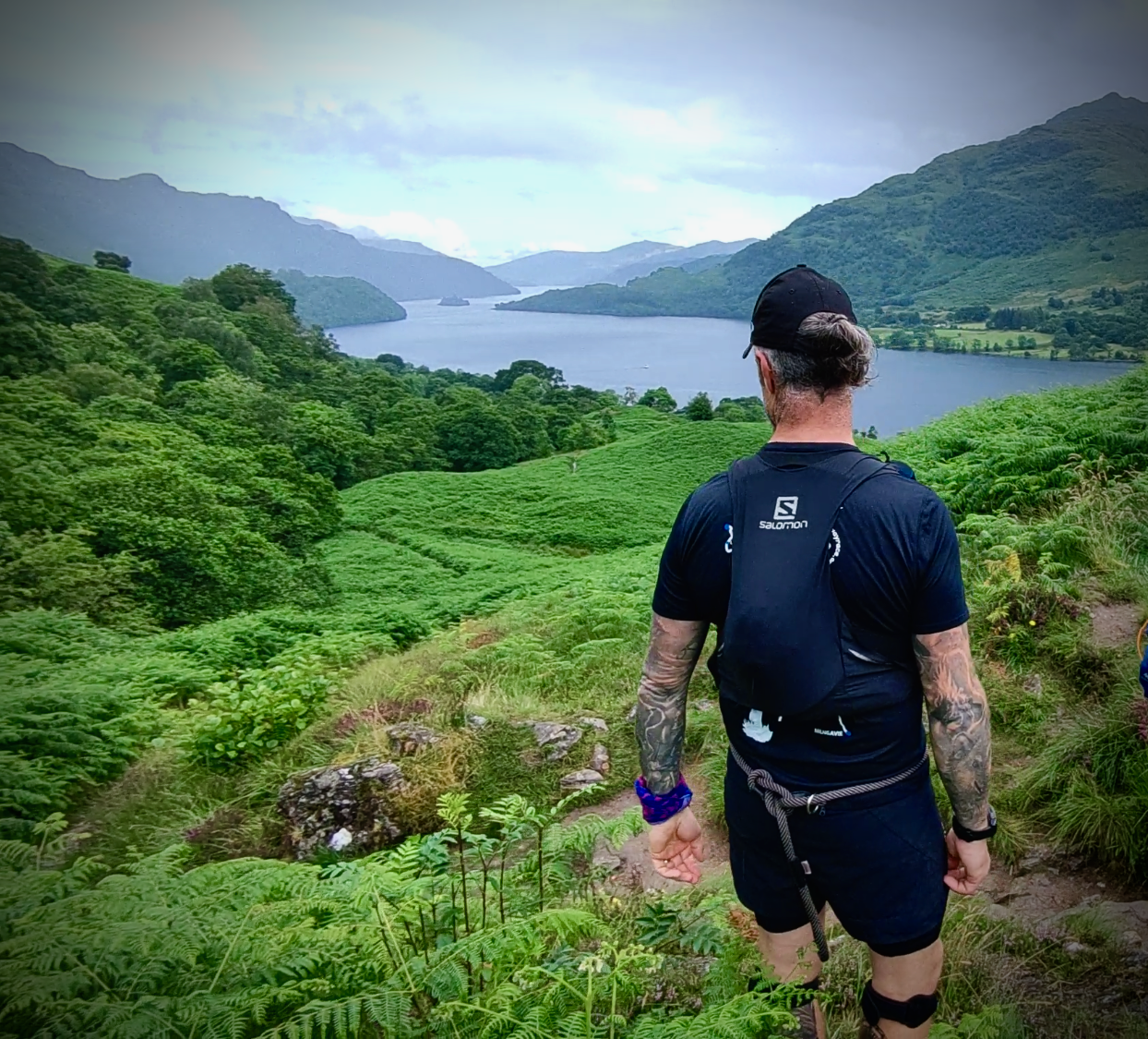 A man with tattoos, wearing a black cap, black shirt, and shorts, carrying a backpack, is hiking on a trail surrounded by lush green vegetation, overlooking a lake and mountains in the distance.