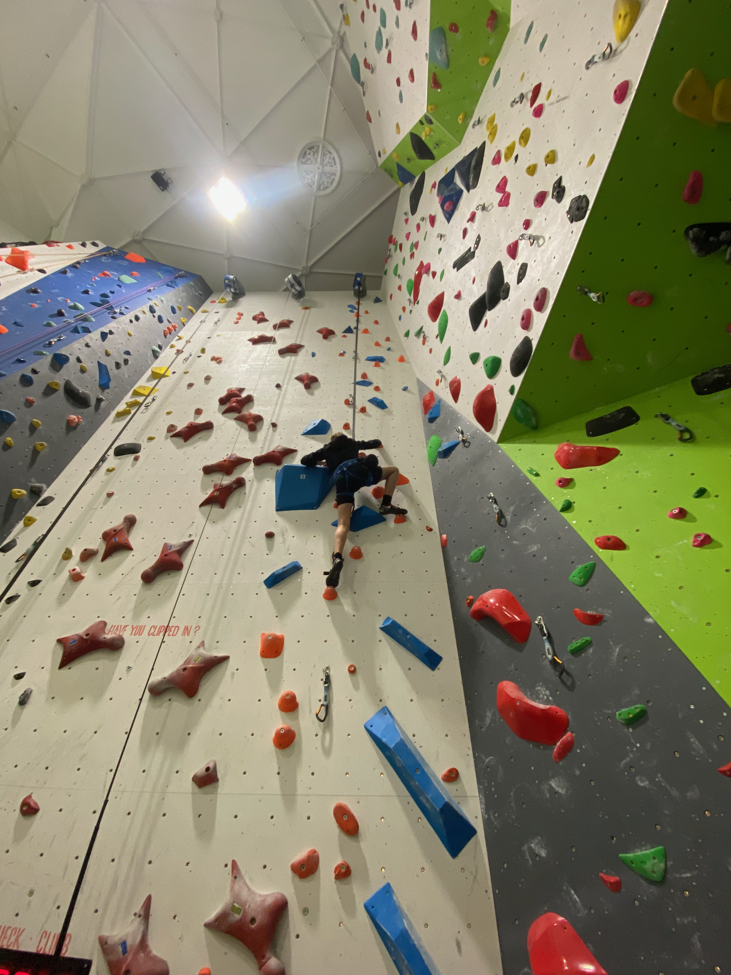 Indoor rock climbing gym with colorful climbing holds on walls of different angles and inclinations. A climber is seen mid-climb, wearing climbing shoes and a harness.