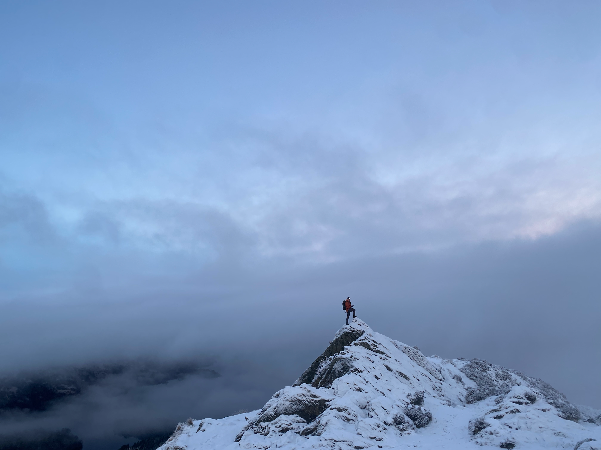 A climber standing on a snow-covered mountain peak holding an ice axe, with a cloudy sky and mountain range in the background.