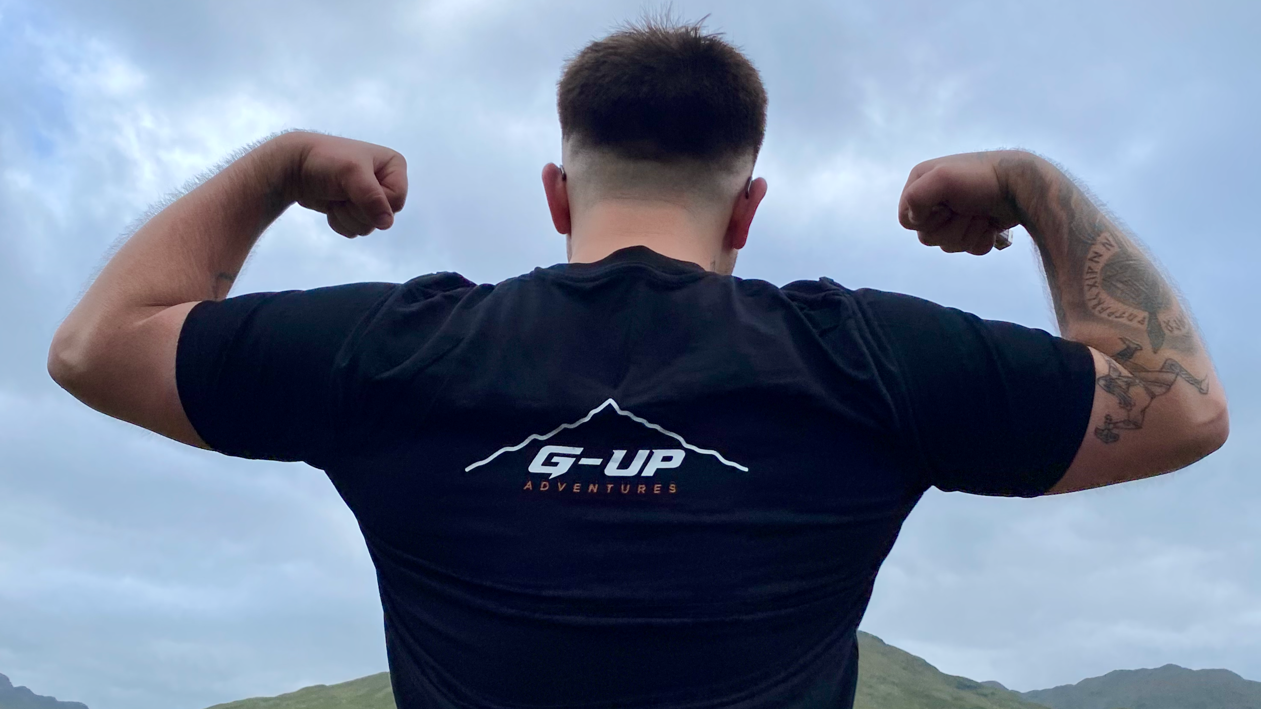 A person flexing their biceps outdoors, seen from behind, with mountains in the distance and a cloudy sky.