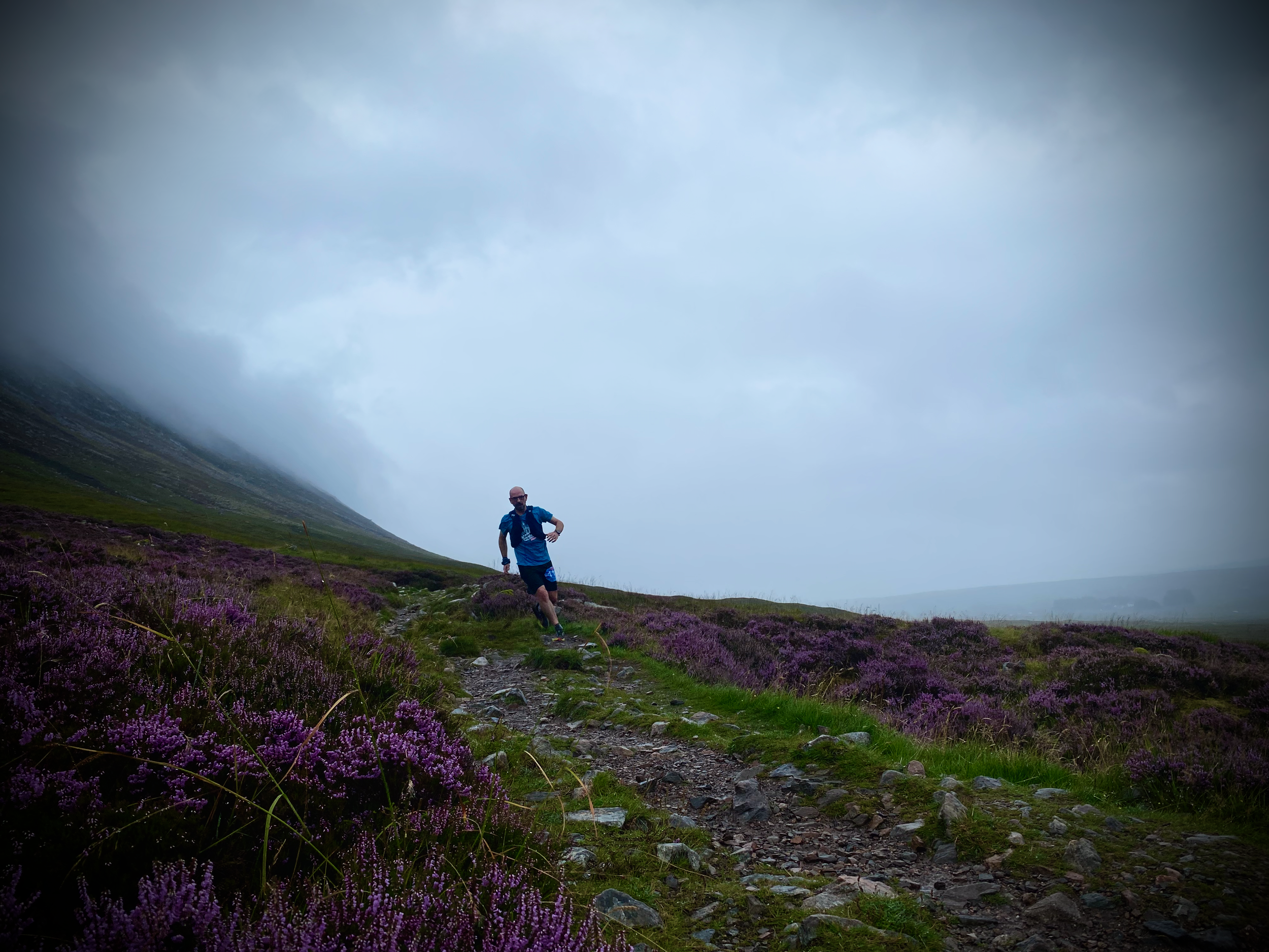 A man hiking on a mountain trail surrounded by purple heather, with cloudy sky and steep hillside in the background.