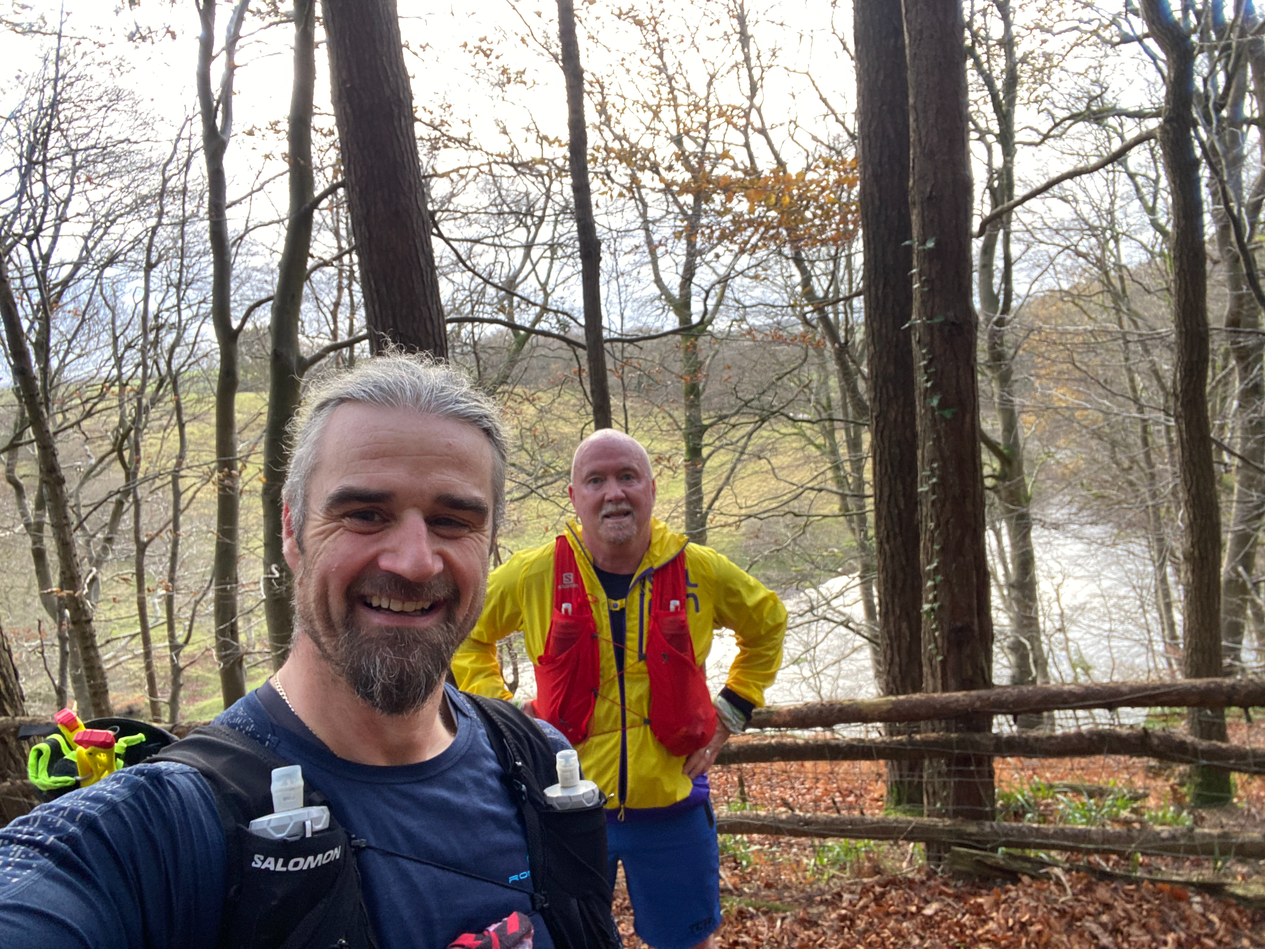 Two male runners or hikers in athletic clothing and gear standing in a forested area with leafless trees and fallen leaves on the ground. Part of a third person's face is visible at the bottom of the image.