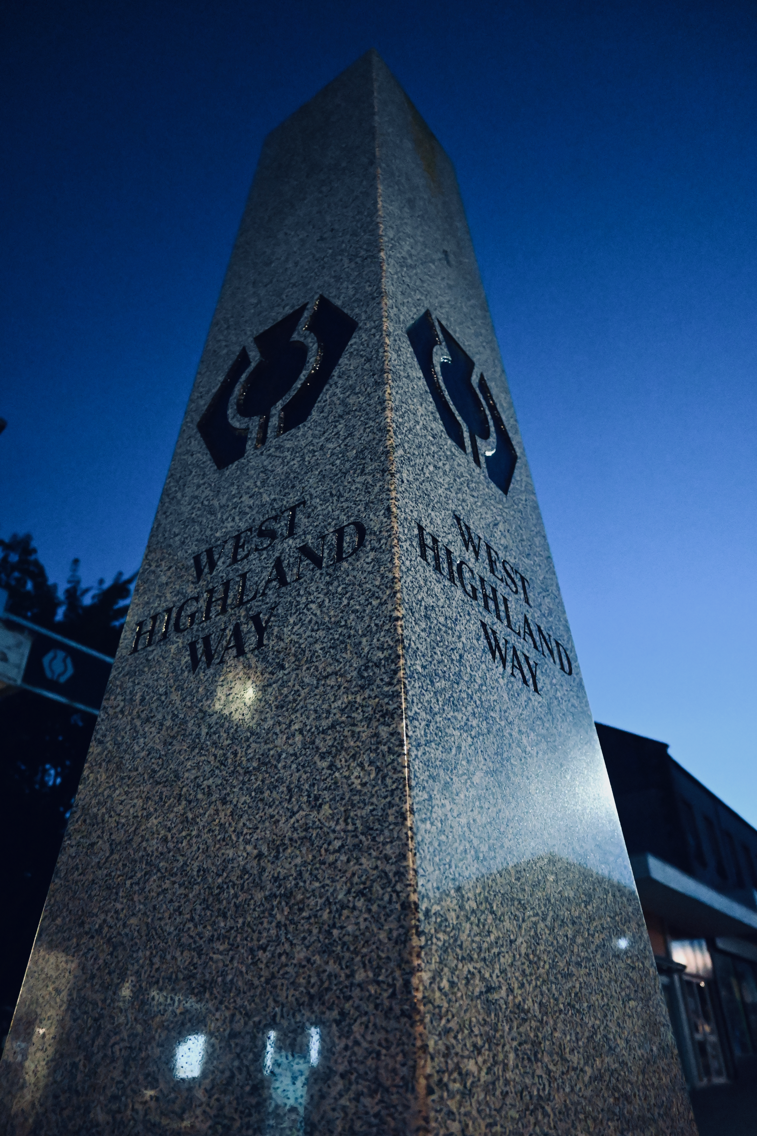 A tall, rectangular granite monument with engraved text and symbols, standing outdoors at dusk against a deep blue sky.