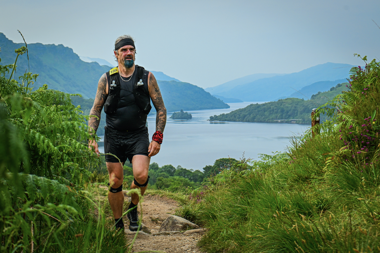 A man running on a trail in a lush, green landscape with a large lake and mountains in the background.
