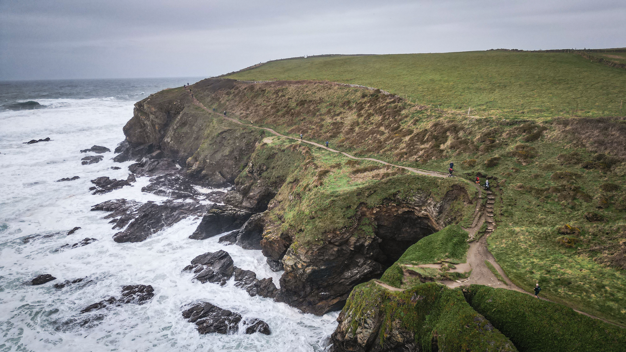 Cliffs along the coast with a narrow trail where several people are walking, overlooking the ocean with waves crashing against the rocks below.