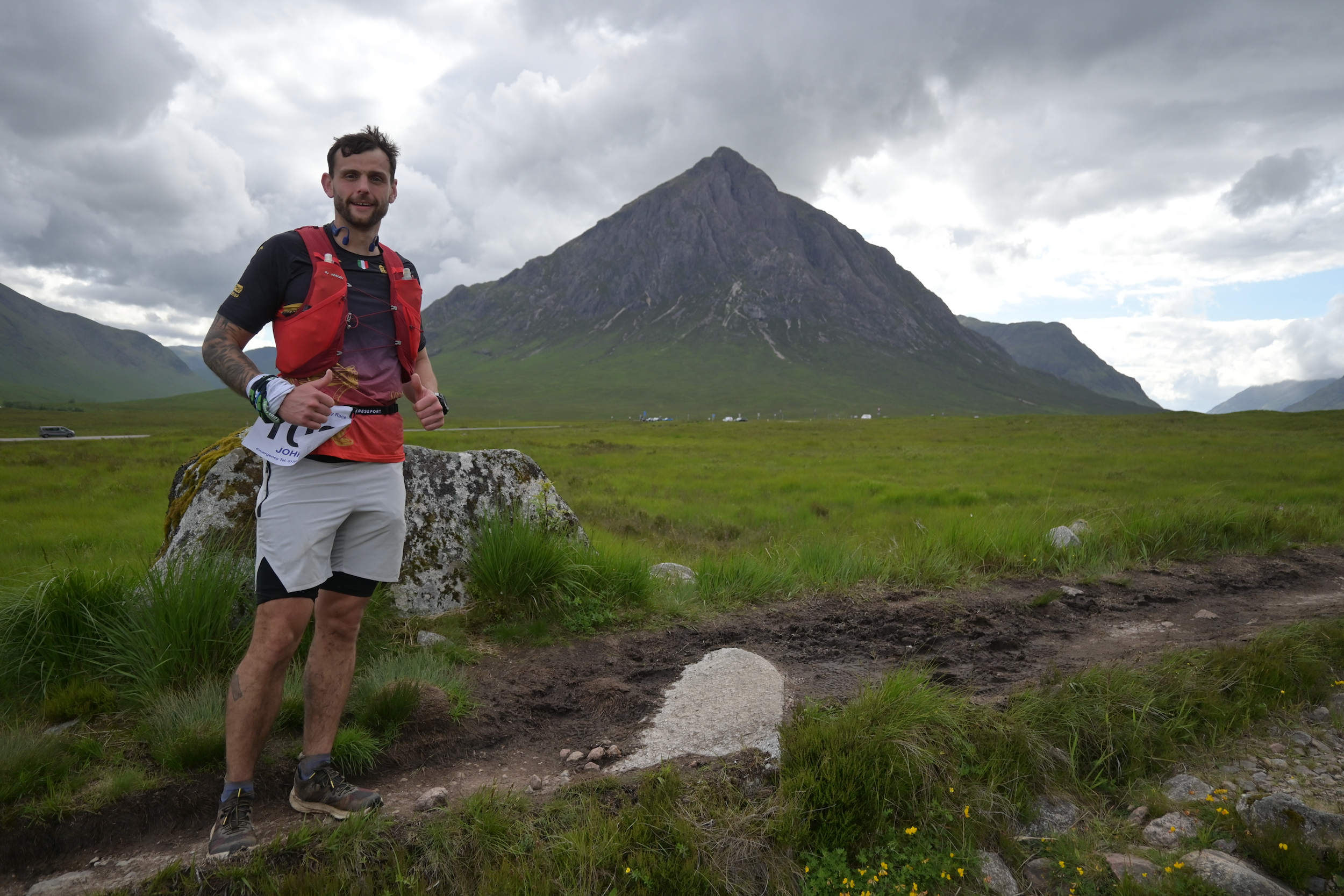 A man with a beard, wearing a black and red running shirt and gray athletic shorts, stands outdoors giving a thumbs up. He is carrying a red hydration pack and has race bib number 107 attached to his shorts. Behind him is a large green mountain under cloudy skies, and the landscape is grassy with some rocks and wildflowers.