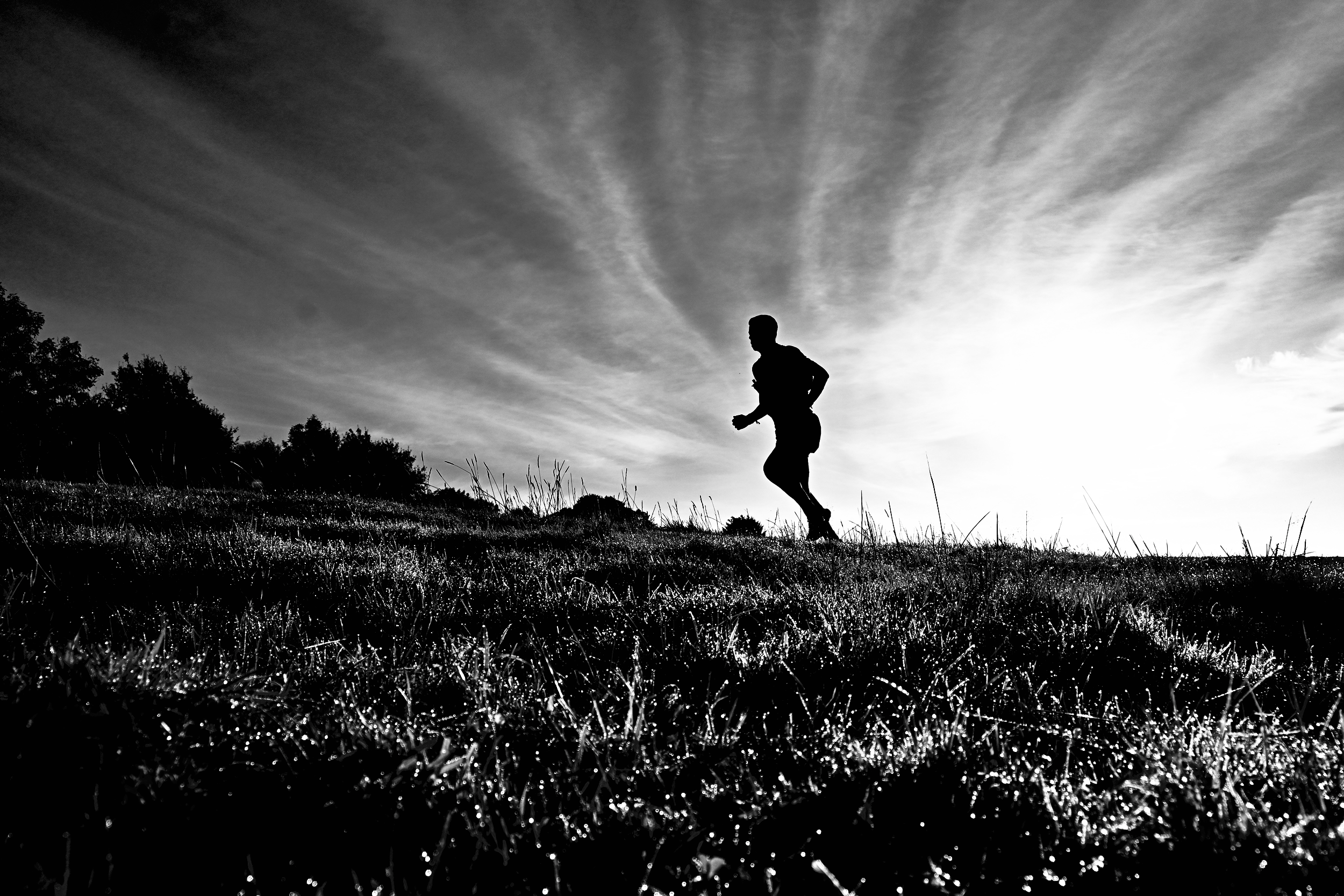 Silhouette of a person running on a grassy hill during sunset with a cloudy sky in the background in black and white.