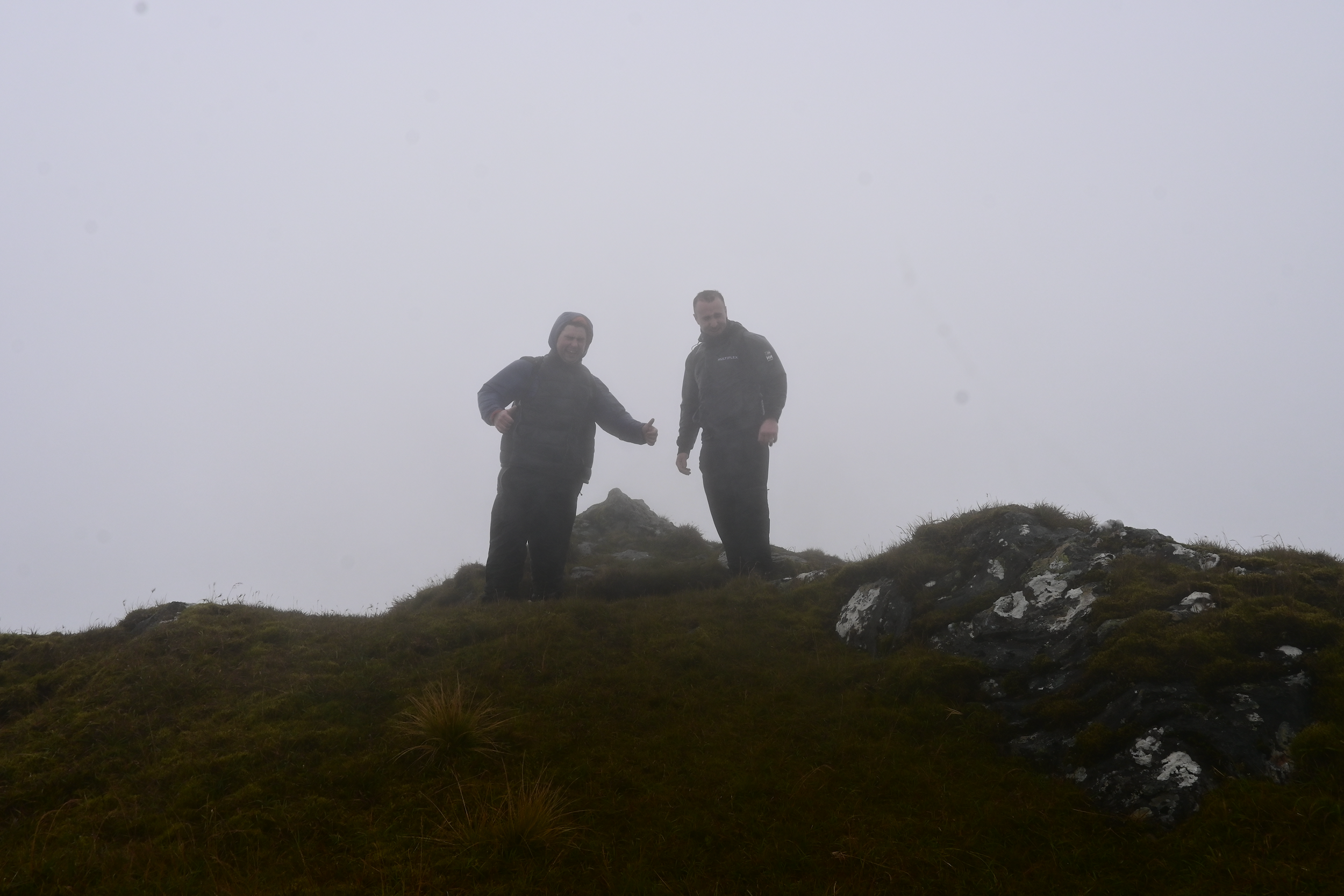 Two men standing on a grassy, rocky hilltop shrouded in fog, giving thumbs-up signs.